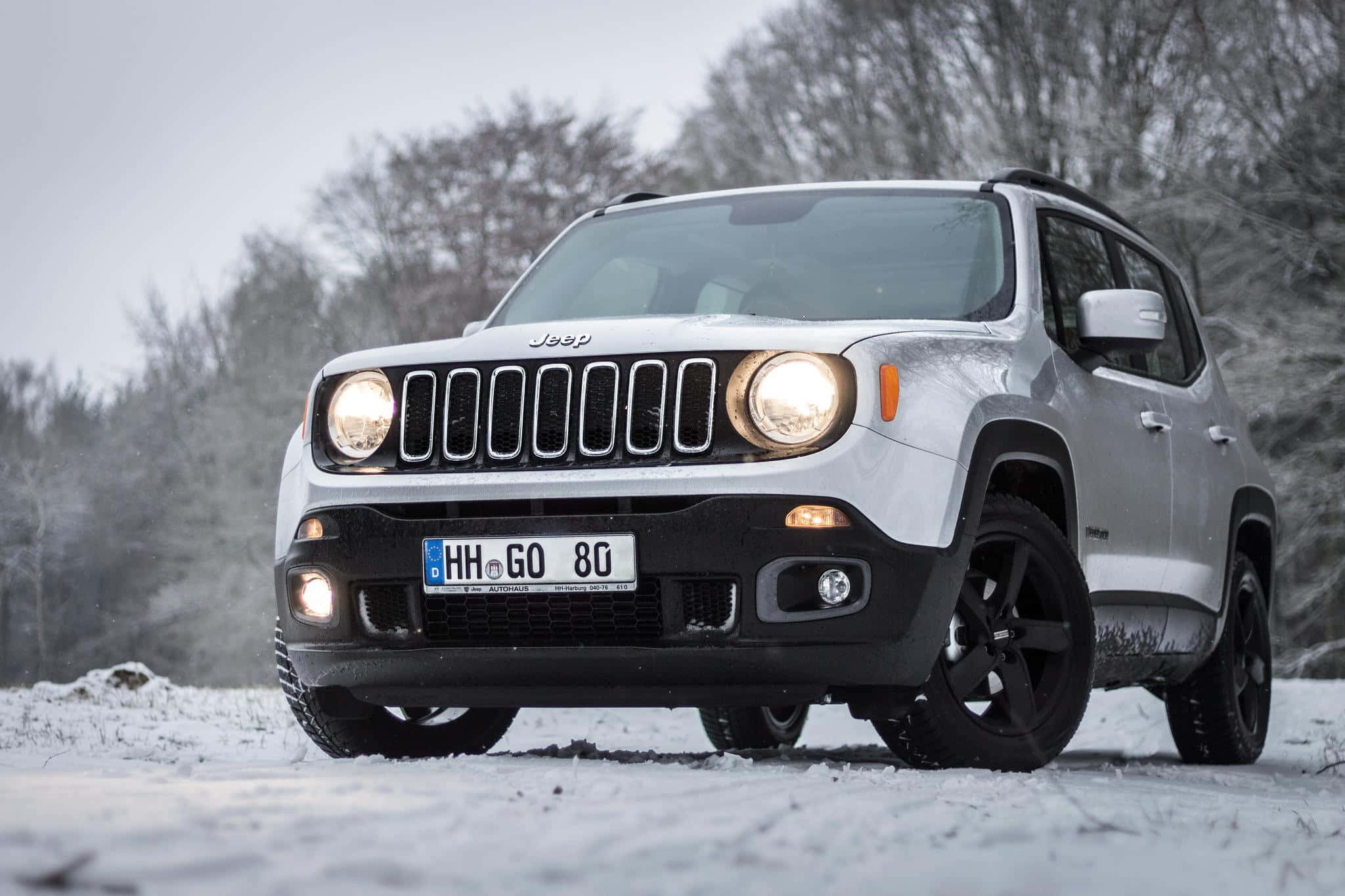 Jeep Renegade 桌布 壁紙 — 一輛白色Jeep Renegade SUV亮著車頭燈，停在雪地上，周圍是光禿禿的冬季樹木，天空多雲。