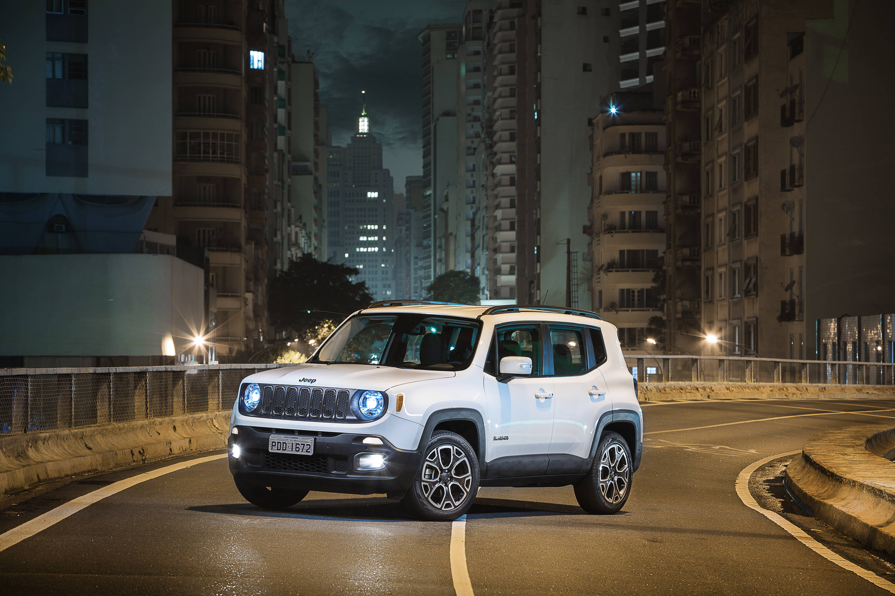 吉普 桌布 壁紙 — 一輛白色Jeep Renegade在夜間行駛於濕漉漉的城市街道上，車頭燈亮起，背景是高聳的建築物。