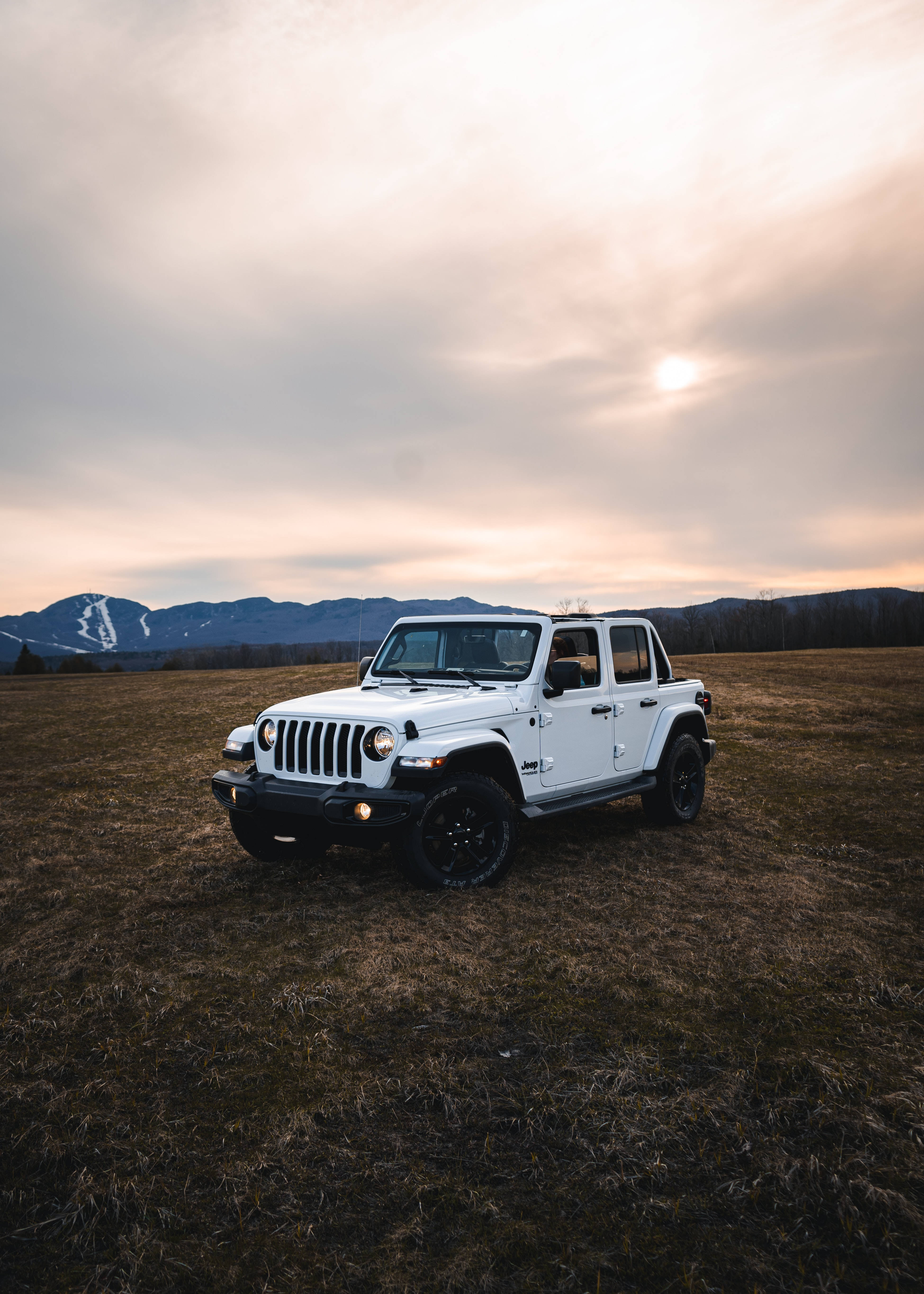 吉普 桌布 壁紙 — 一輛白色Jeep Wrangler獨自矗立在廣闊的田野中，夕陽西下，背景是白雪覆蓋的山脈。