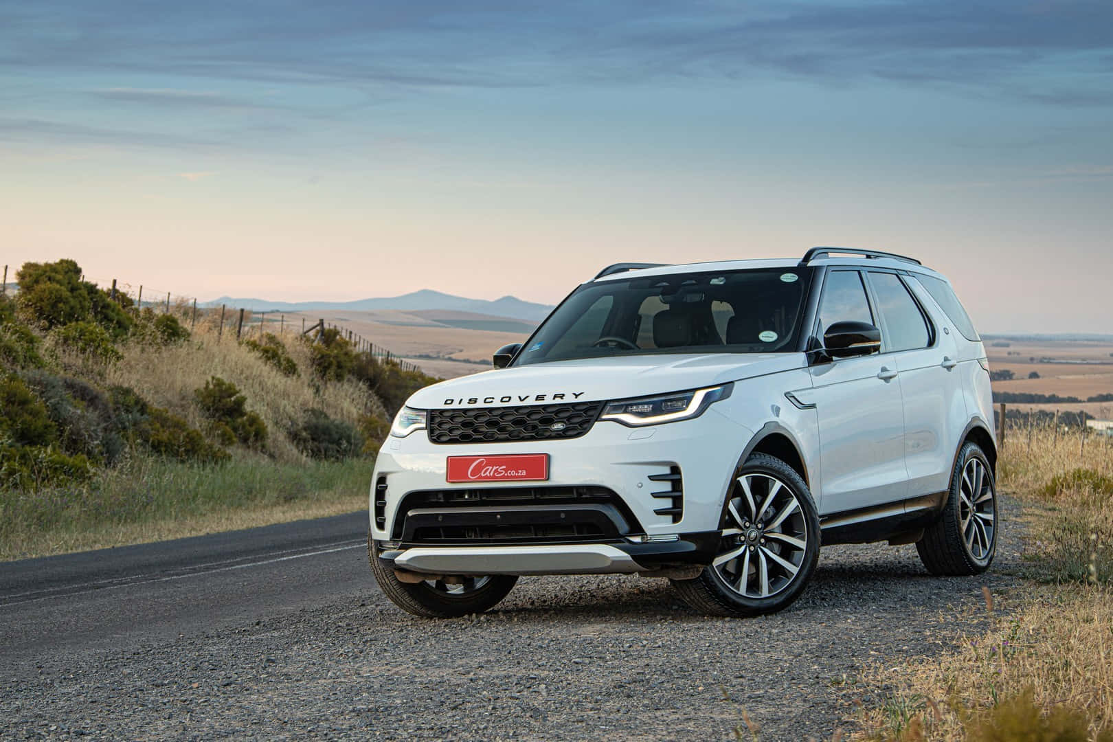 Land Rover Discovery 桌布 壁紙 — 一輛白色Land Rover Discovery在黃昏時停泊在碎石路上，背景是起伏的山丘和乾草。