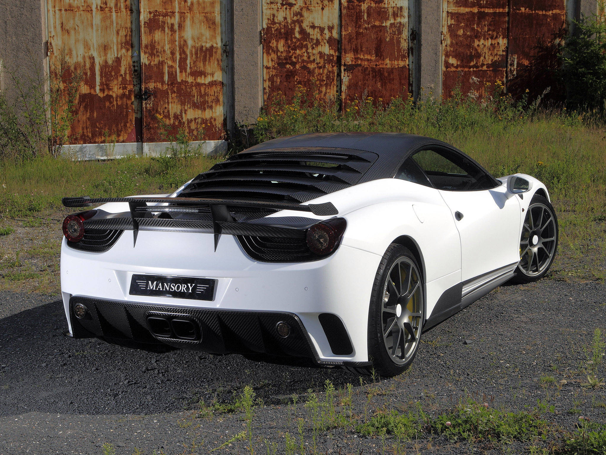 white ferrari 桌布 壁紙 — 一輛白色Mansory改裝法拉利跑車，帶有碳纖維飾邊，停在碎石路上，前面是一扇生鏽的金屬門。