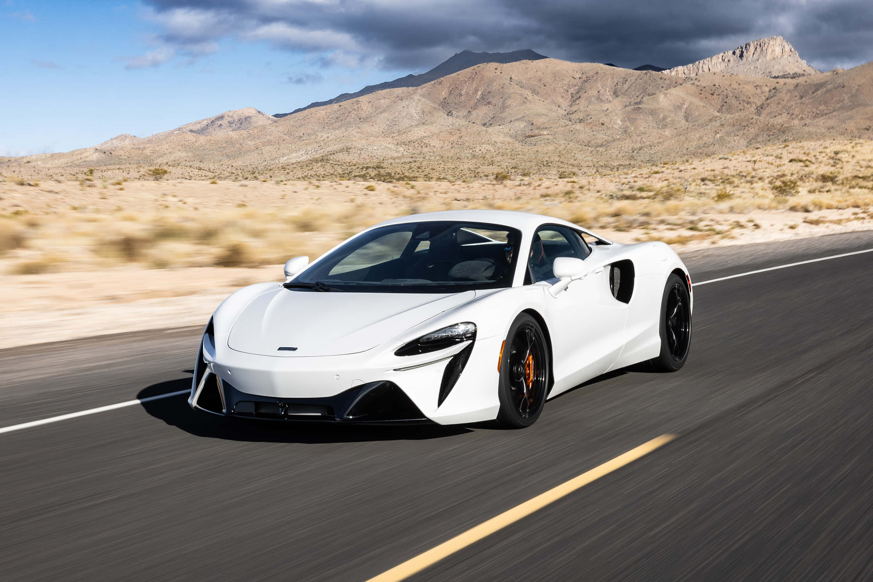 麥拉倫 桌布 壁紙 — 一輛白色McLaren跑車在部分多雲的天空下，沿著崎嶇山脈的沙漠高速公路飛馳。