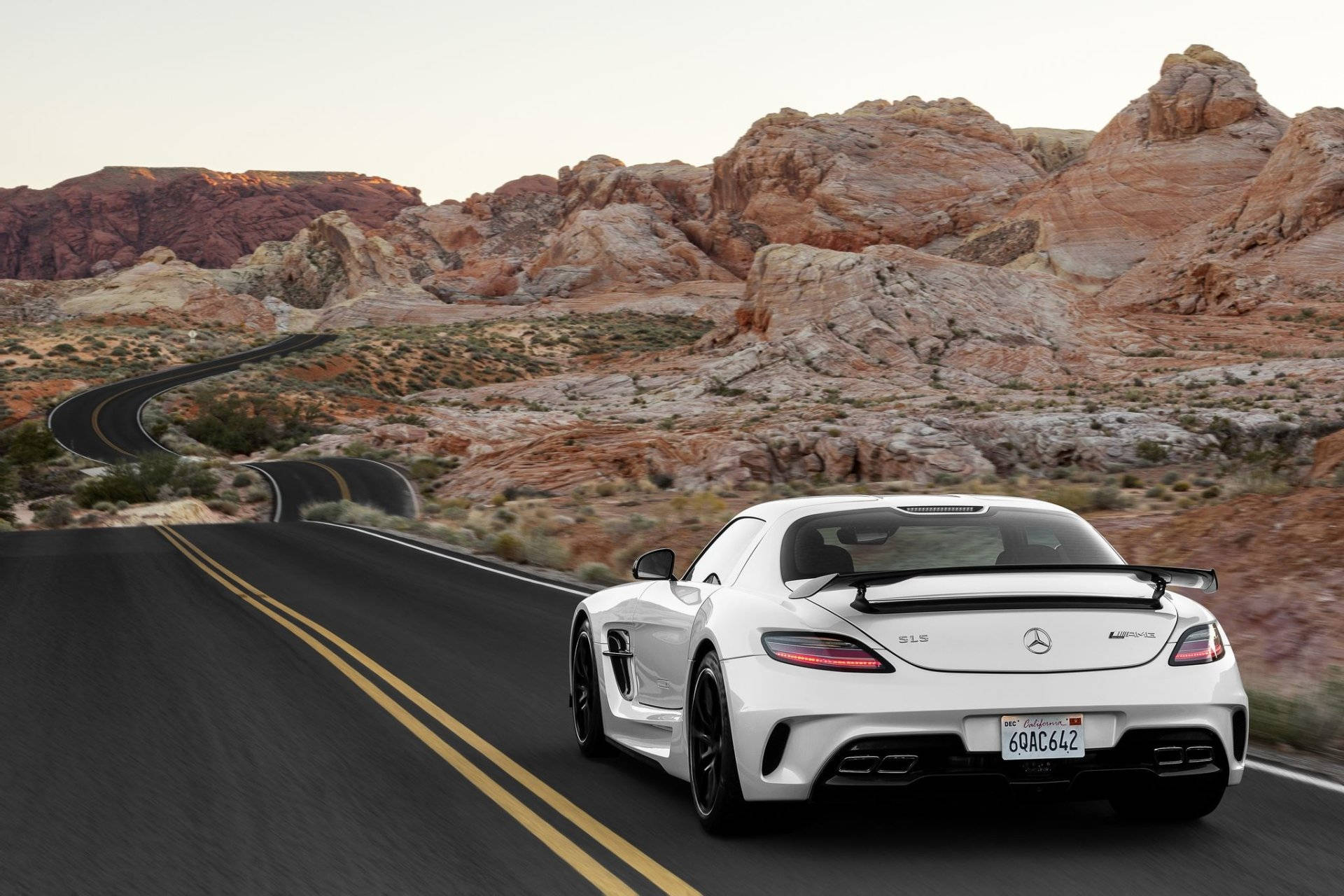 Mercedes Benz Amg 桌布 壁紙 — 一輛白色Mercedes-AMG SLS GT3跑車在日落時分沿著蜿蜒的沙漠公路疾馳，兩旁是紅色岩層。