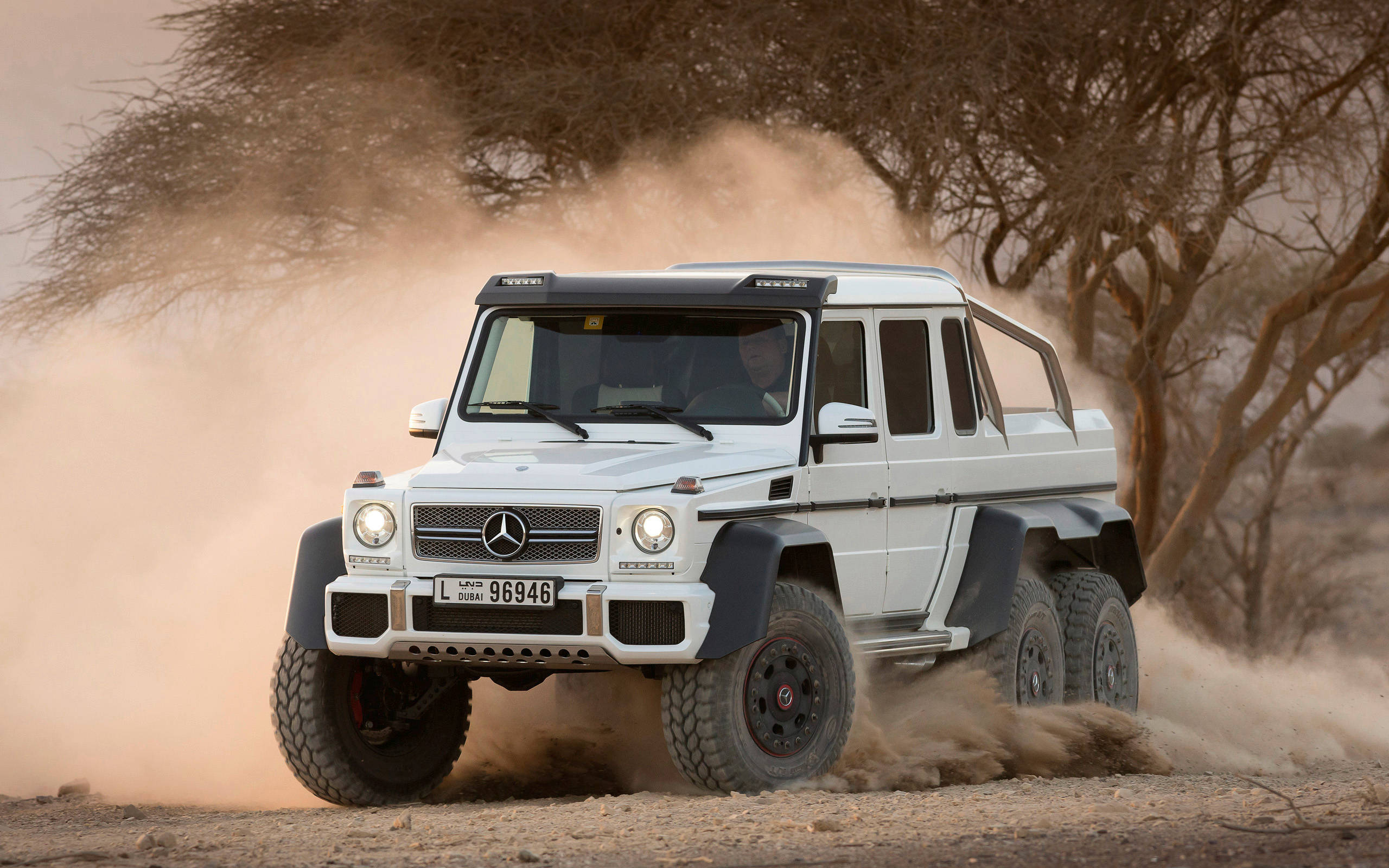 amg iPhone 桌布 壁紙 — 一輛白色Mercedes-Benz G63 6x6裝甲車在乾燥的稀樹草原上疾馳，揚起塵土，稀疏的金合歡樹點綴其間。