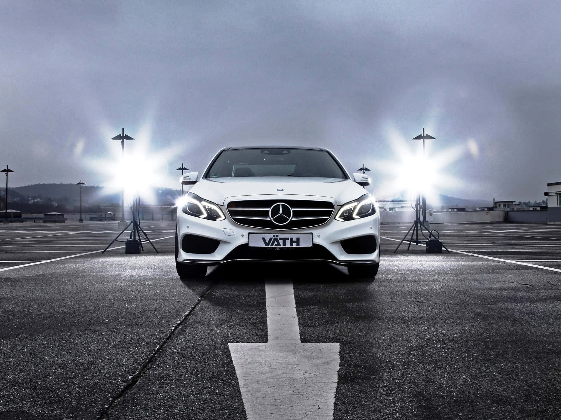 Car Front View 桌布 壁紙 — 一輛白色Mercedes房車在潮濕的停車場上向前，兩旁有明亮的攝影棚燈光，天空多雲。