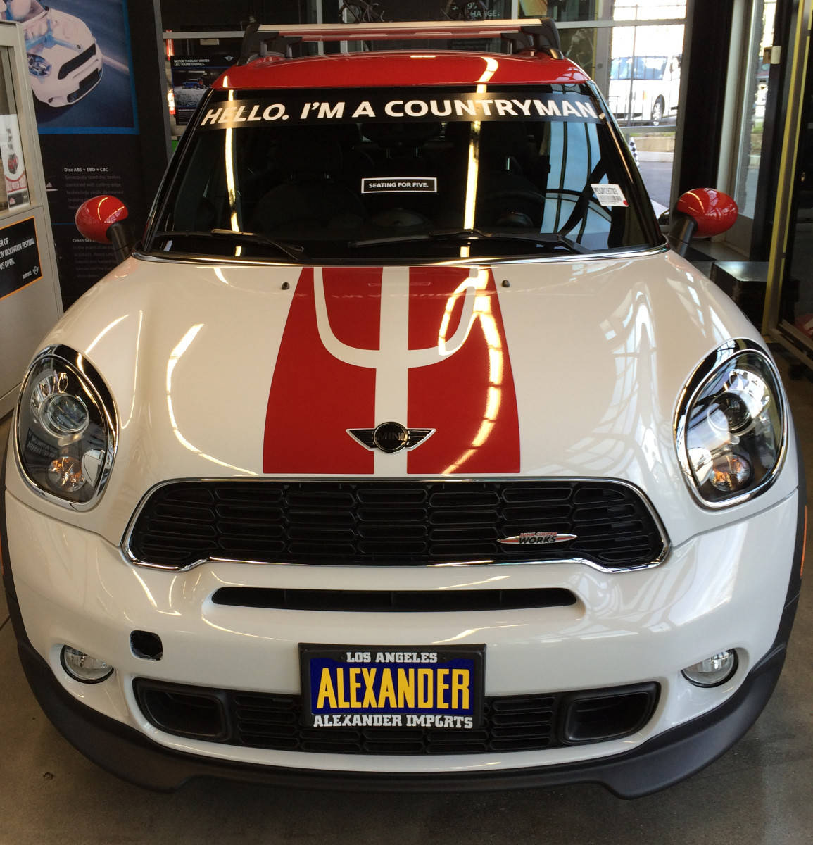 Mini 桌布 壁紙 — 一輛白色MINI Countryman，帶有紅色賽車條紋，擋風玻璃上寫著「HELLO I'M A COUNTRYMAN」，停放在展廳內。