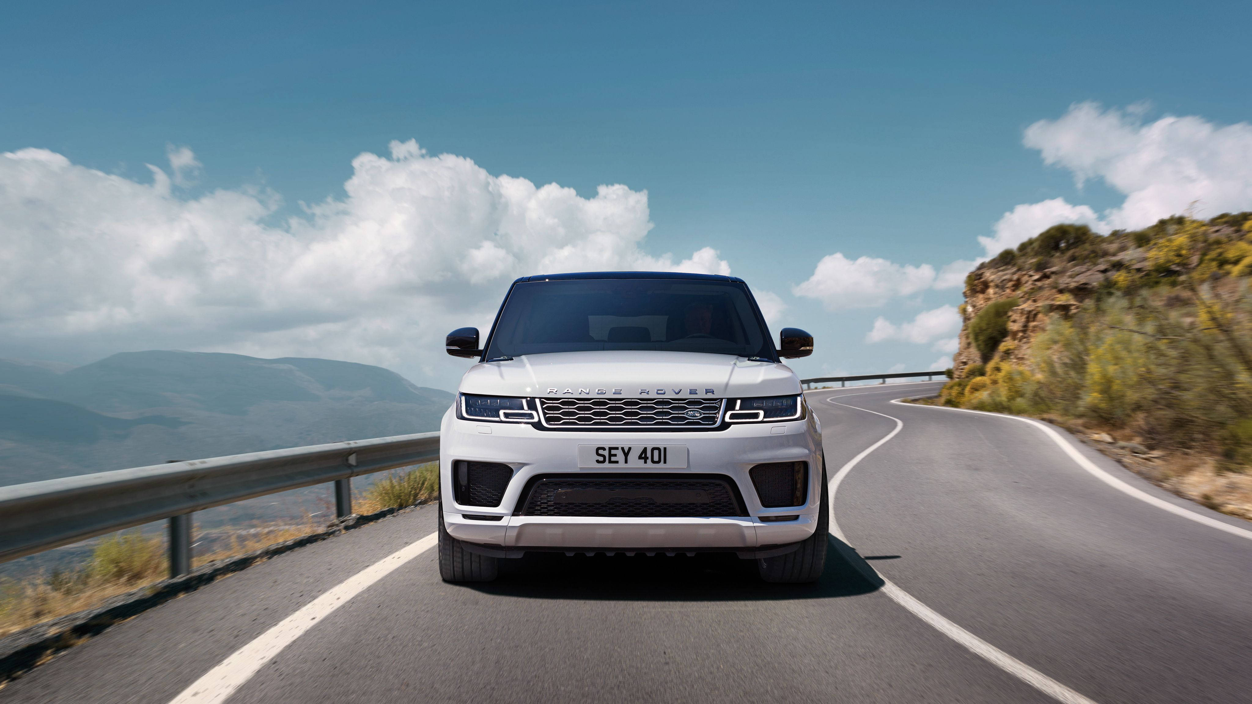 荒原路華 桌布 壁紙 — 一輛白色Range Rover Sport在晴朗藍天和蓬鬆白雲下，自信地行駛在蜿蜒的山路上。