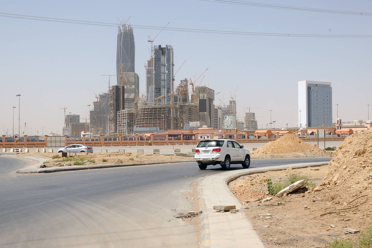Riyadh 桌布 壁紙 — 一輛白色SUV在彎曲的道路上行駛，經過正在開發的建築工地，高聳的摩天大樓在朦朧的天空下。