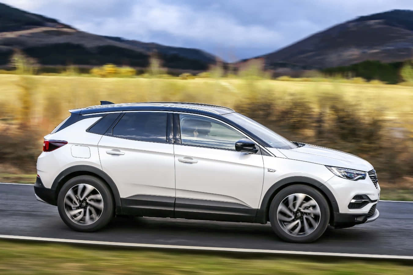 Opel Grandland X 桌布 壁紙 — 一輛白色SUV在陰天之下沿著蜿蜒的道路穿過起伏的丘陵疾馳。