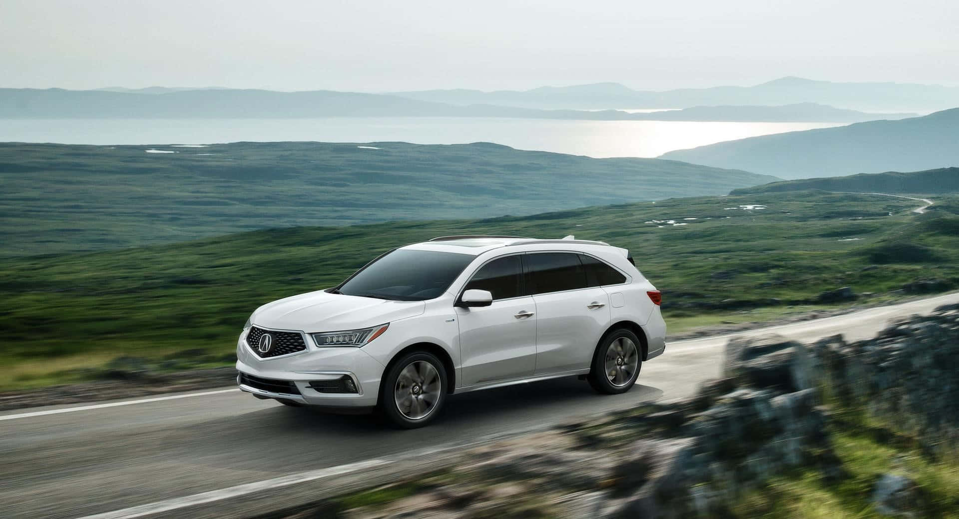 Acura Mdx 桌布 壁紙 — 一輛白色SUV沿著蜿蜒的海岸道路行駛，兩旁是鬱鬱蔥蔥的綠色山丘，遠處是波光粼粼的大海，天空朦朧。