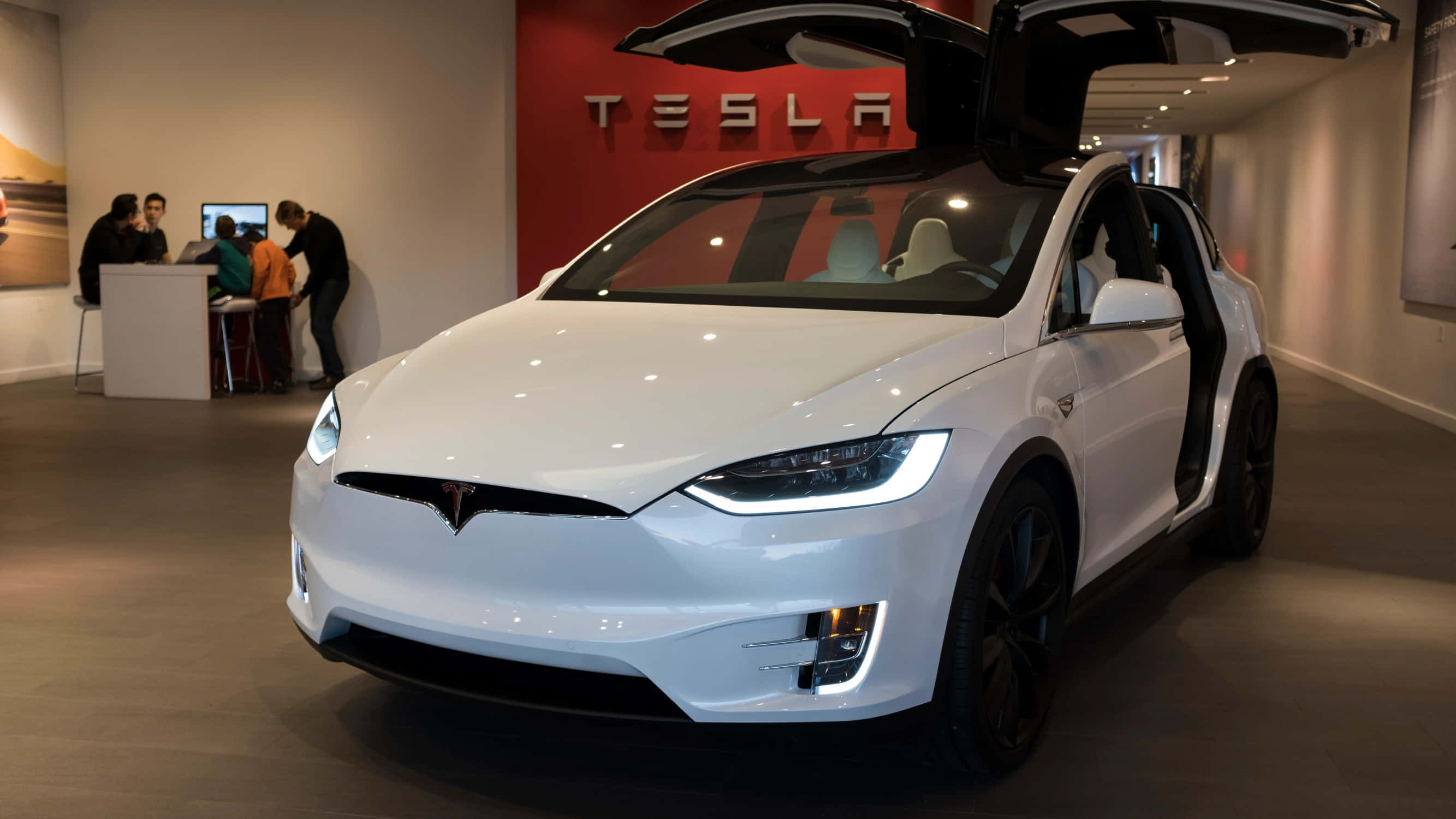 特斯拉 桌布 壁紙 — 一輛白色Tesla Model X打開鷹翼門，在現代化展廳中展示，背景有紅色品牌標識和顧客。