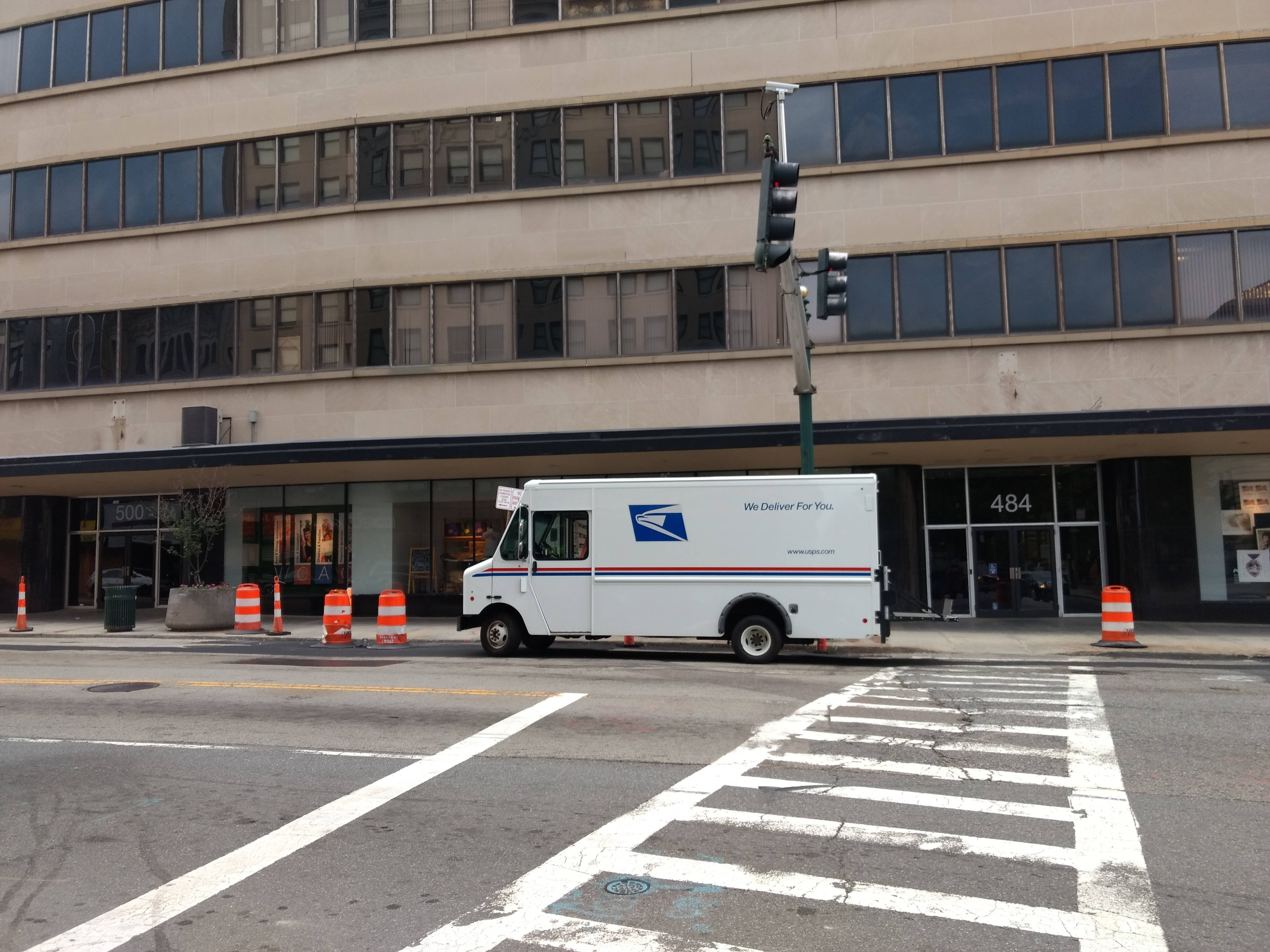 Truck 桌布 壁紙 — 一輛白色USPS送貨車停在城市街角，附近有米色辦公大樓、交通號誌和橙色三角錐。