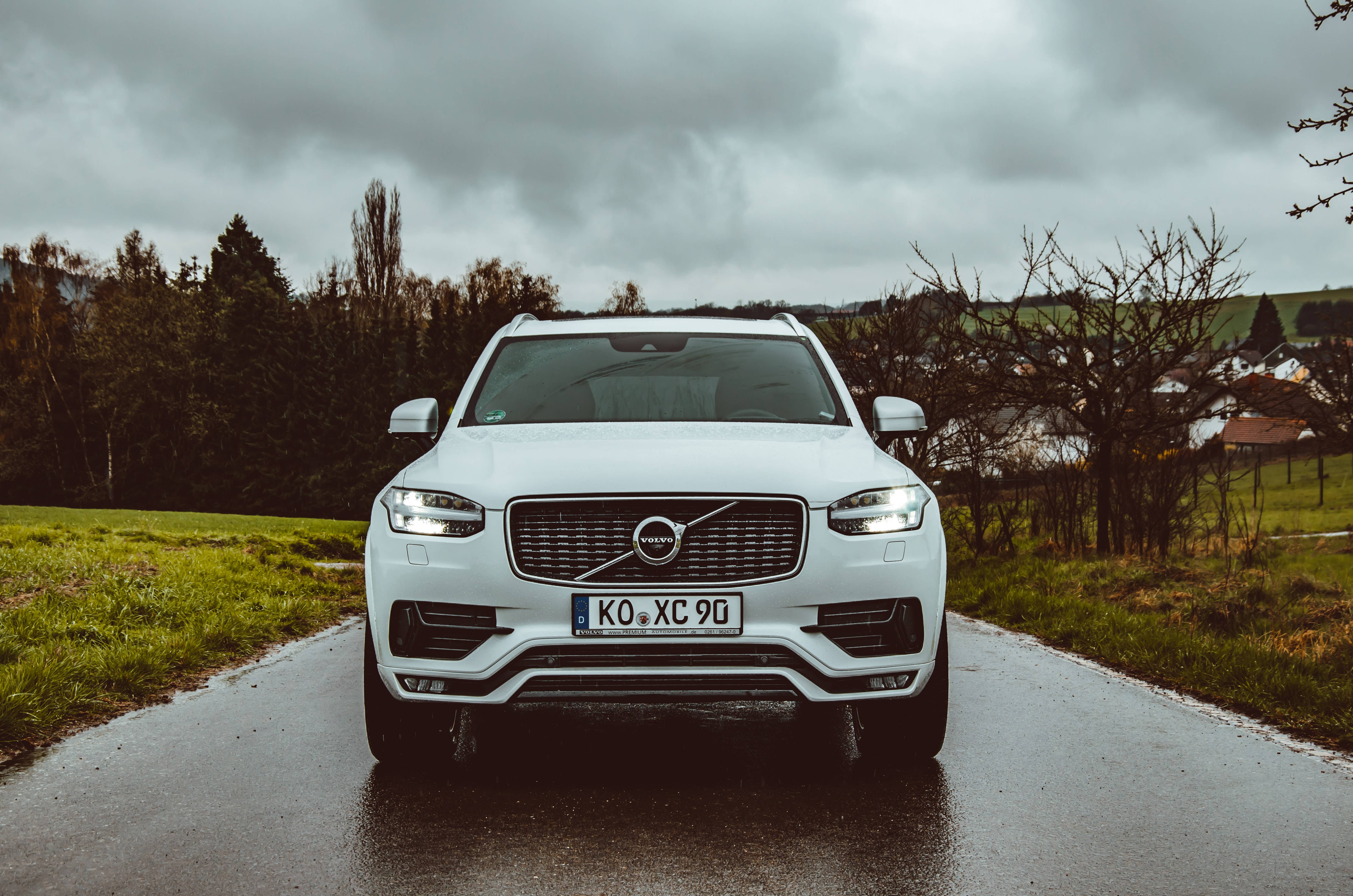Volvo 桌布 壁紙 — 一輛白色Volvo XC90 SUV在潮濕的鄉村道路上向前行駛，天空多雲，背景有綠色田野和光禿禿的樹木。