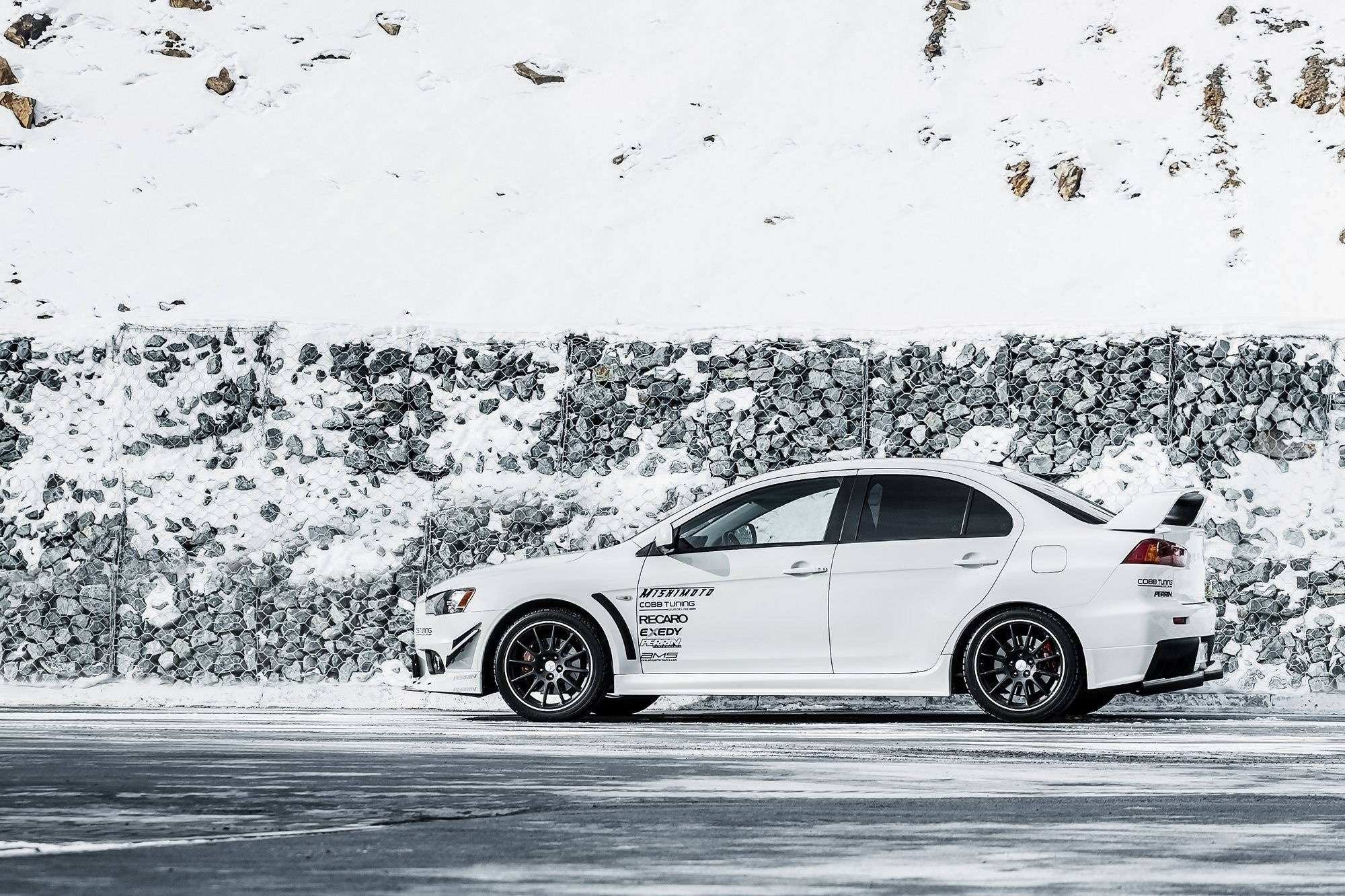 三菱 桌布 壁紙 — 一輛白色三菱Lancer Evolution跑車在冬天停放在雪路旁的石牆邊。