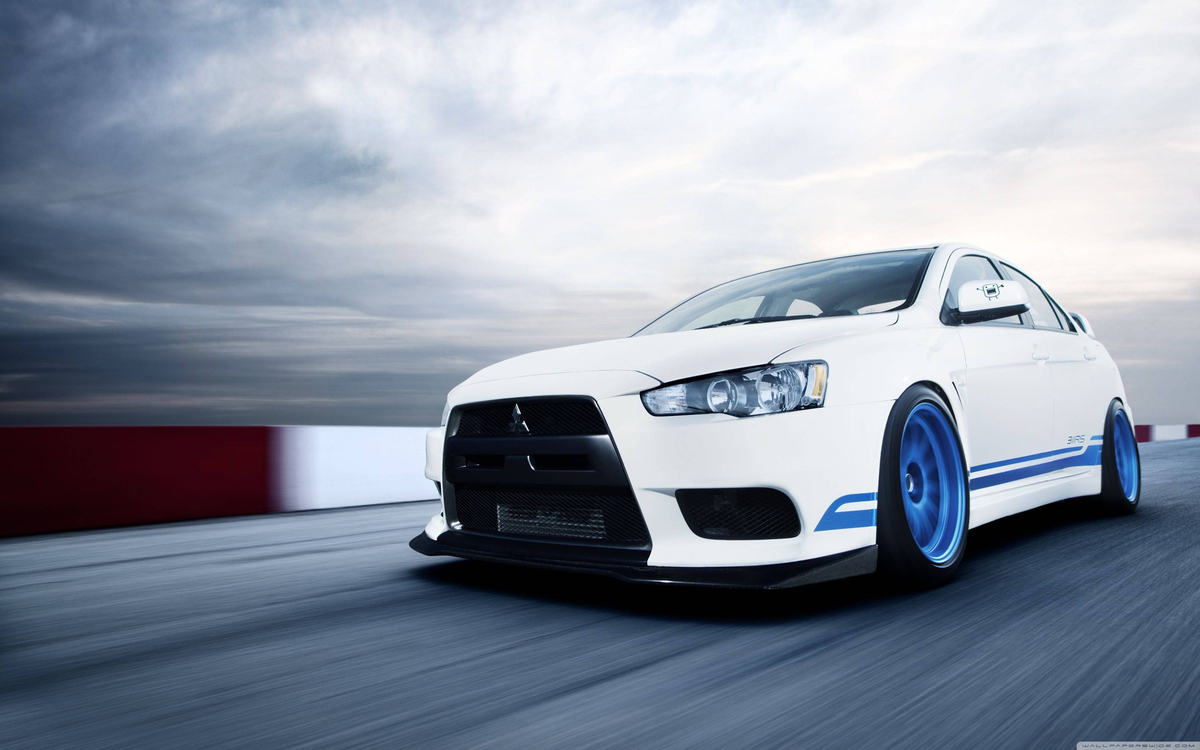 Mitsubishi 桌布 壁紙 — 一輛白色三菱Lancer Evolution搭配藍色輪圈，在多雲的天空下於賽道上疾馳。