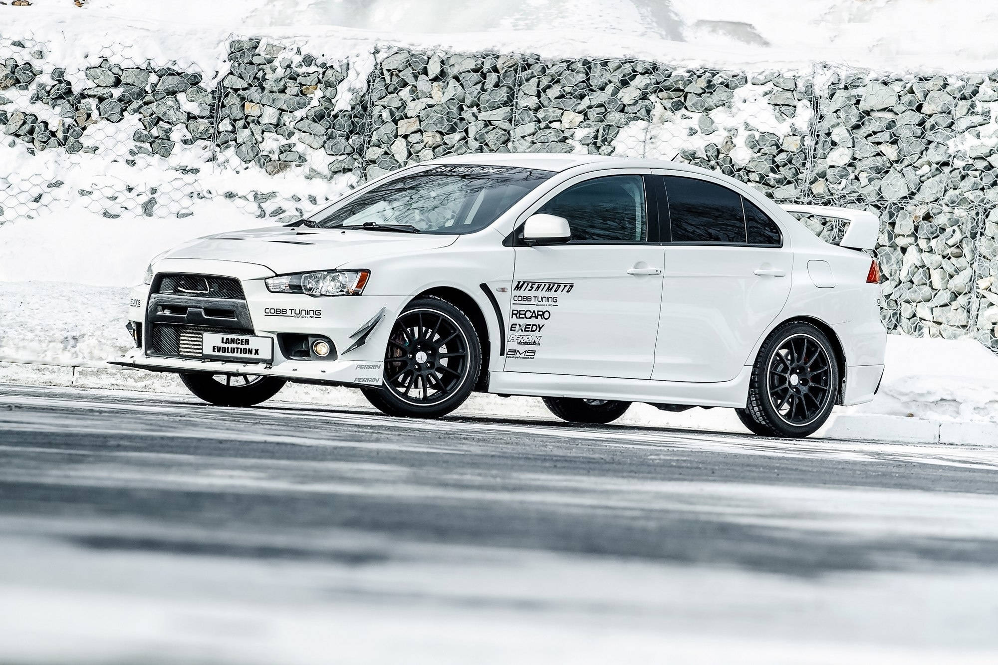 三菱 桌布 壁紙 — 一輛白色三菱Lancer Evolution X在明亮的日光下，在石牆旁邊的雪路上飄移。