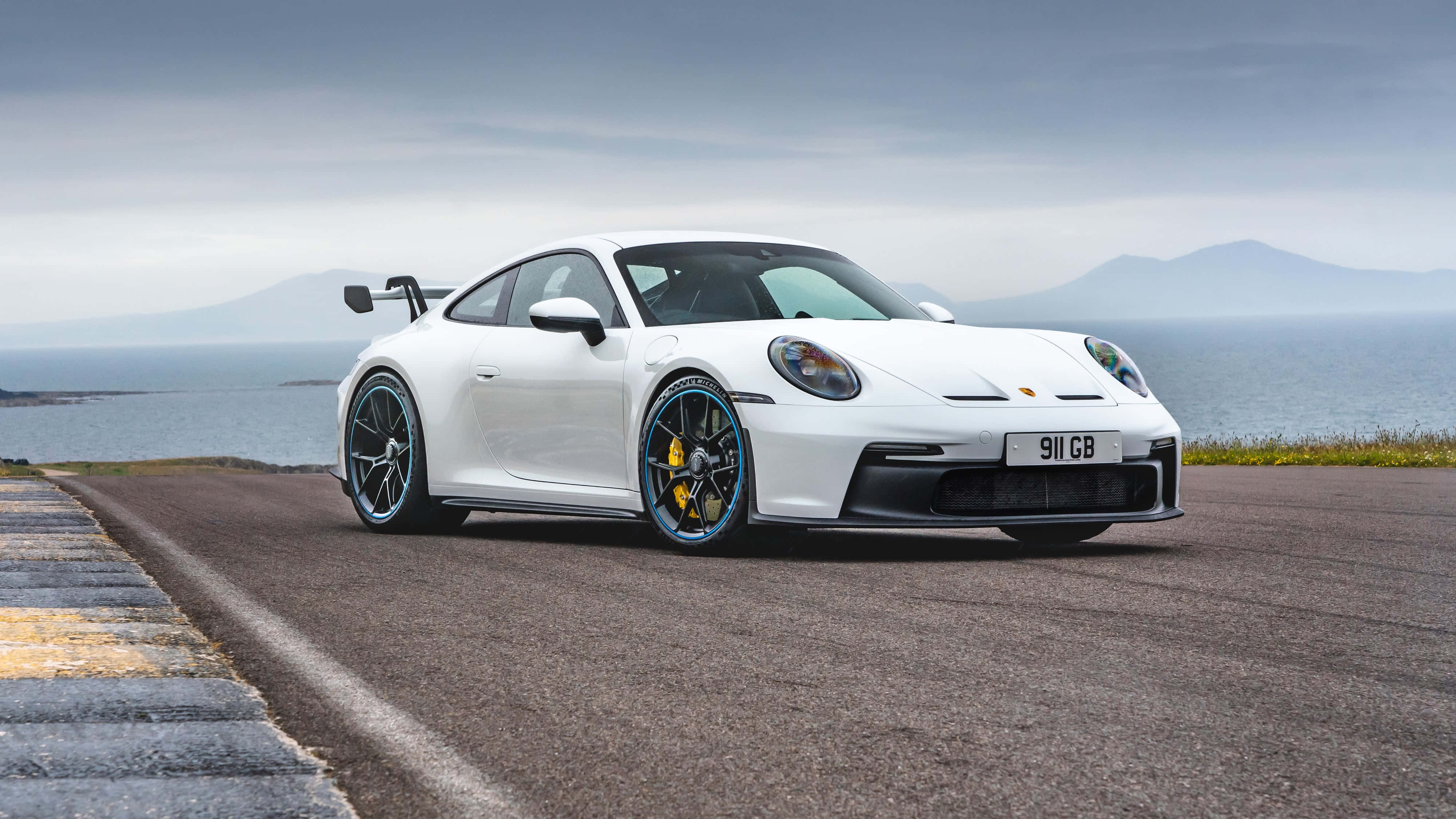 桌布 壁紙 — 一輛白色保時捷911 GT3 RS，帶有藍色飾邊和黃色制動卡鉗，停泊在俯瞰大海的海岸公路上。