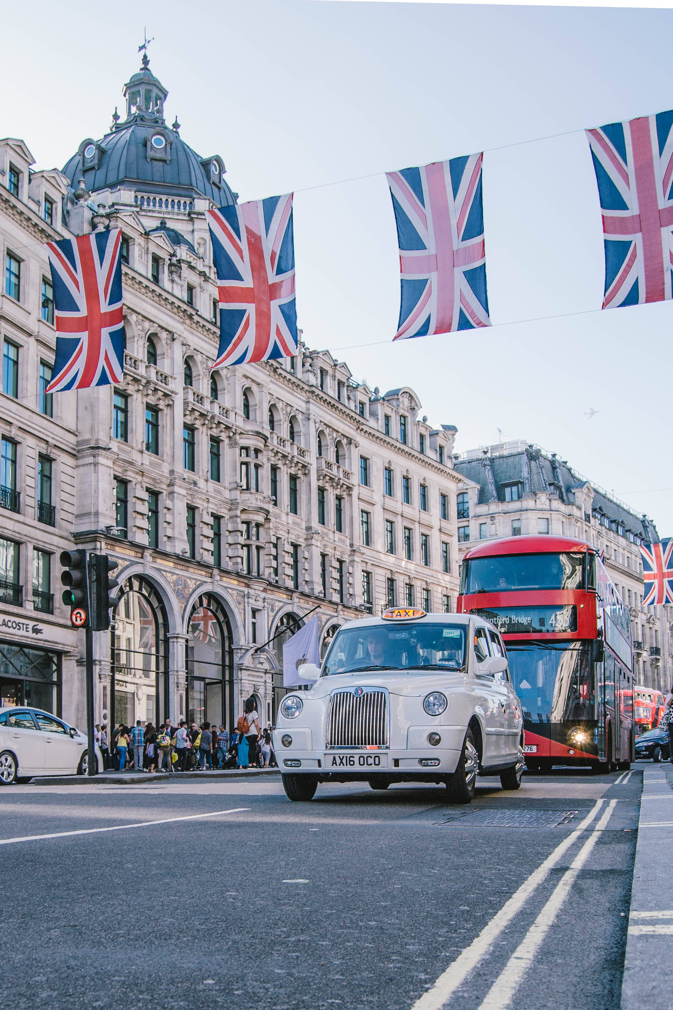 United Kingdom Flag 桌布 壁紙 — 一輛白色倫敦計程車和紅色雙層巴士在懸掛於歷史石造建築上的英國國旗下行駛。