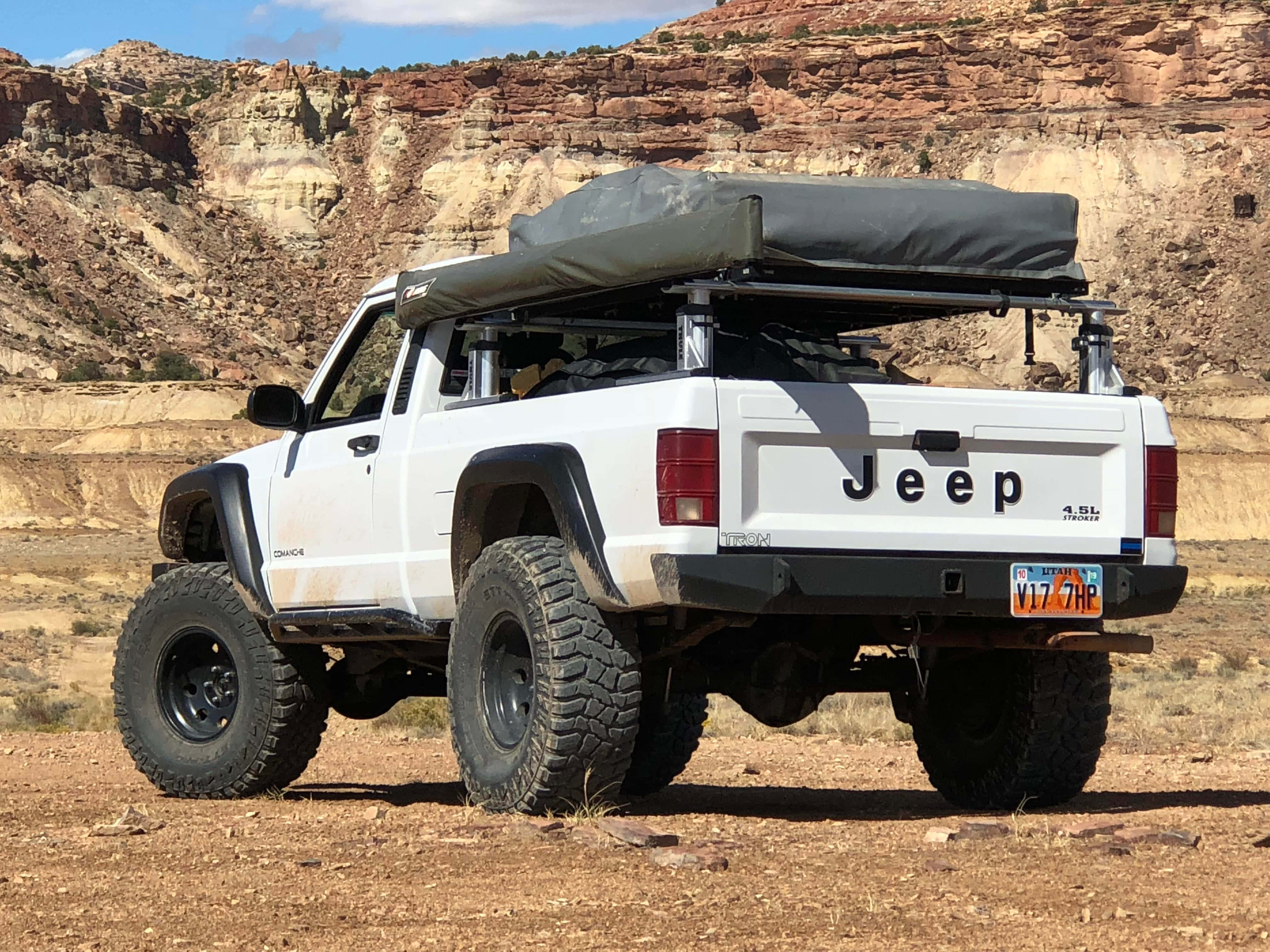 Jeep Comanche 桌布 壁紙 — 一輛白色升高底盤的Jeep Wrangler，車頂帳篷，停在紅土上，背景是層層疊疊的峽谷岩層，天空湛藍明亮。