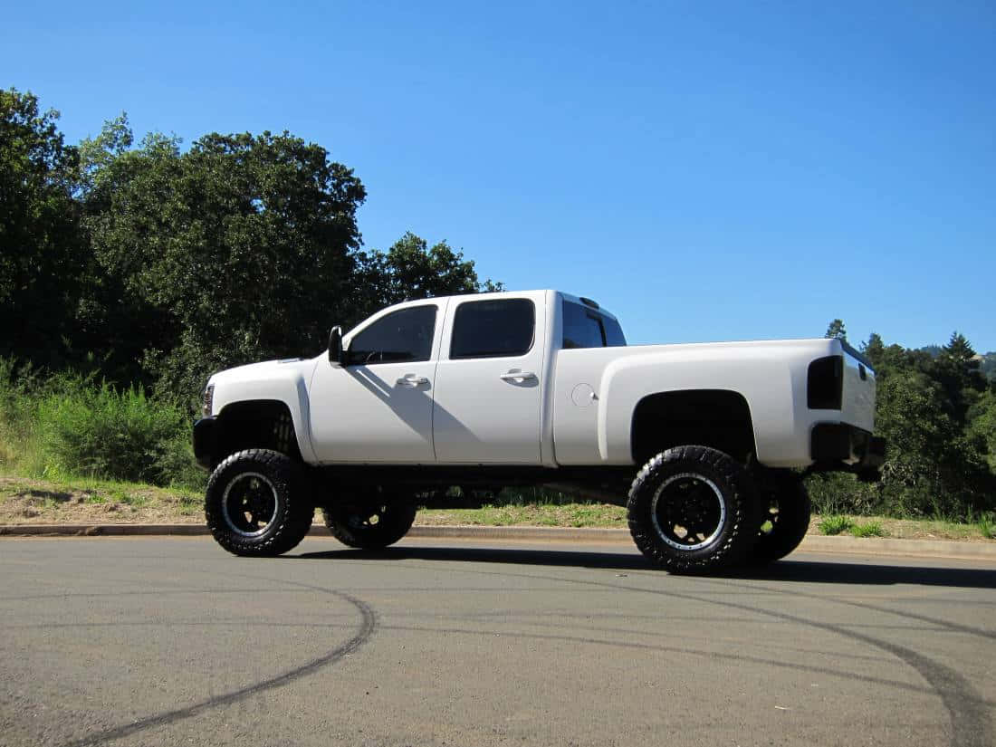 Jacked Up Trucks 桌布 壁紙 — 一輛白色升高版雪佛蘭Silverado皮卡車，配備特大黑色輪胎，停泊在鋪砌道路上，背景是晴朗的藍天和綠樹。