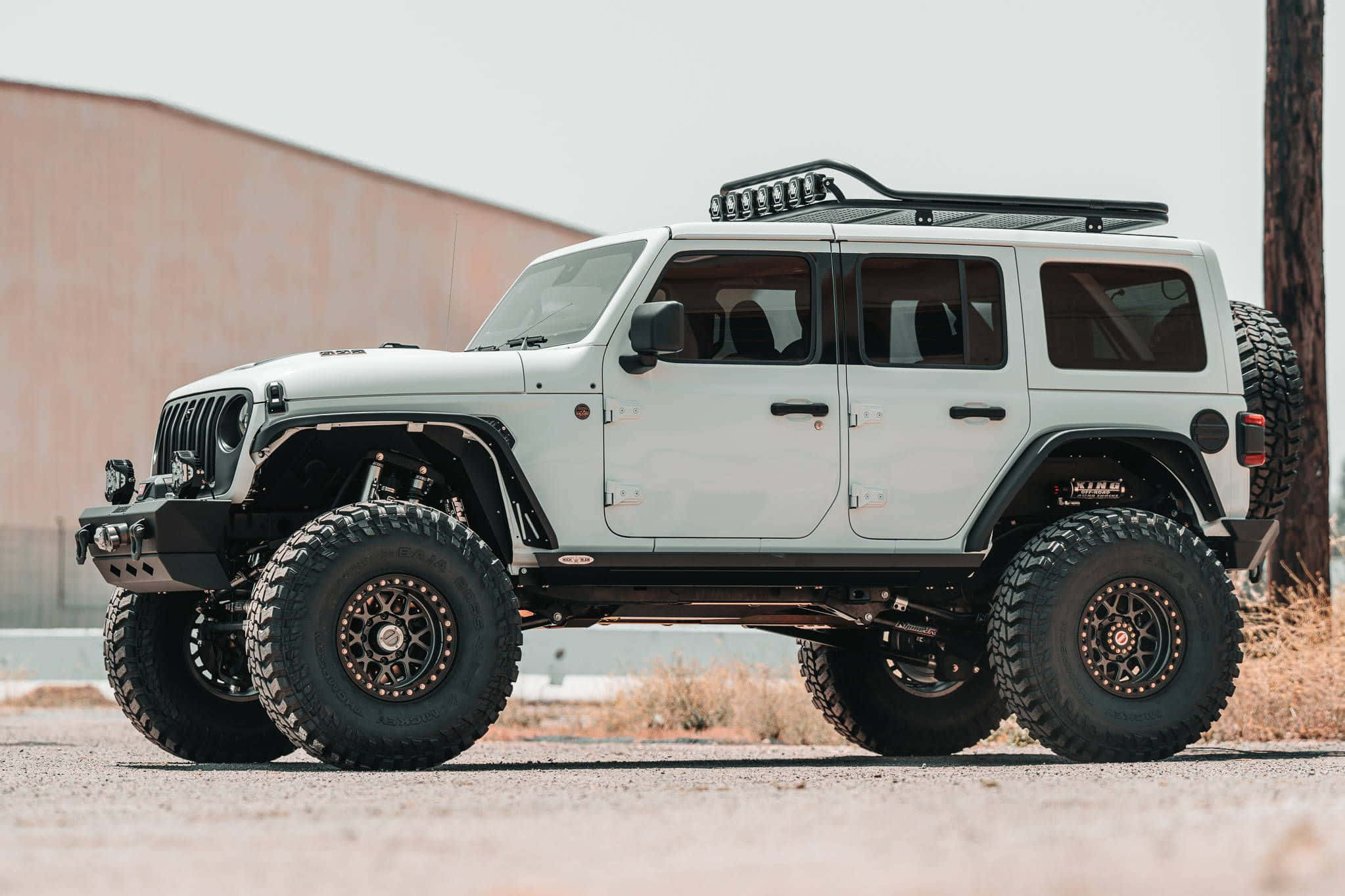Off-road Car 桌布 壁紙 — 一輛白色升高的Jeep Wrangler，配備超大輪胎和車頂行李架，停放在碎石上，附近有木柱和米色牆。