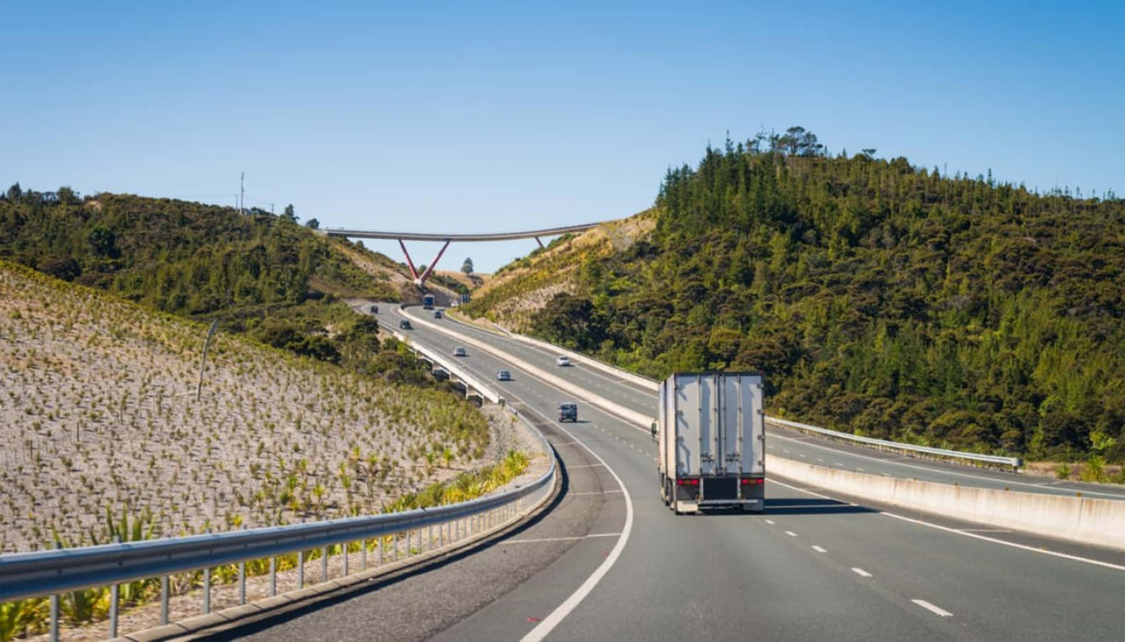 Highways 桌布 壁紙 — 一輛白色卡車在蜿蜒的高速公路上行駛，穿過陽光明媚的山丘，上方有一座現代橋樑在清澈的藍天下延伸。