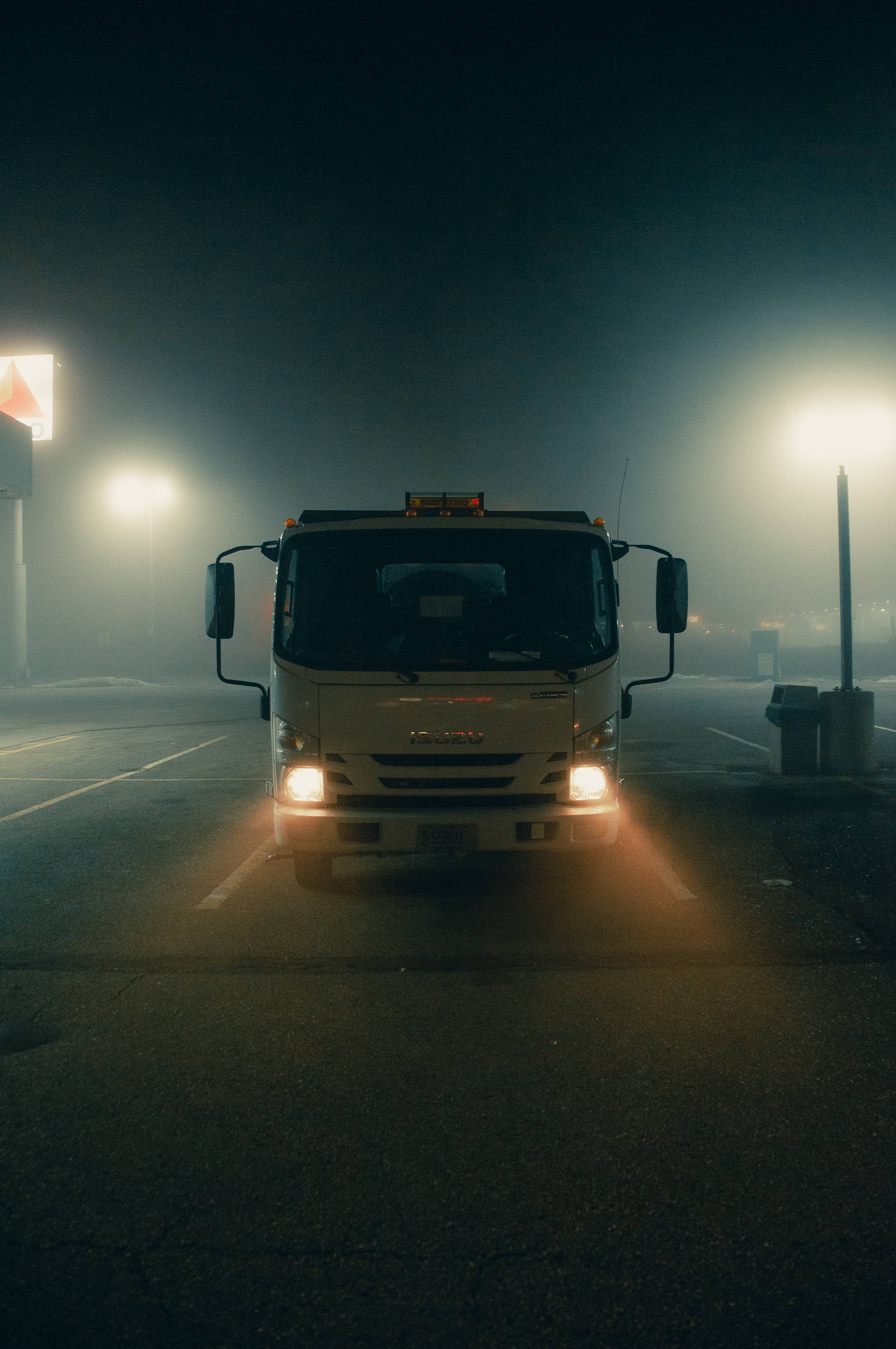 卡車 桌布 壁紙 — 一輛白色卡車的車頭燈在夜間穿透濃霧，照亮昏暗的停車場，遠處的路燈閃爍著光芒。