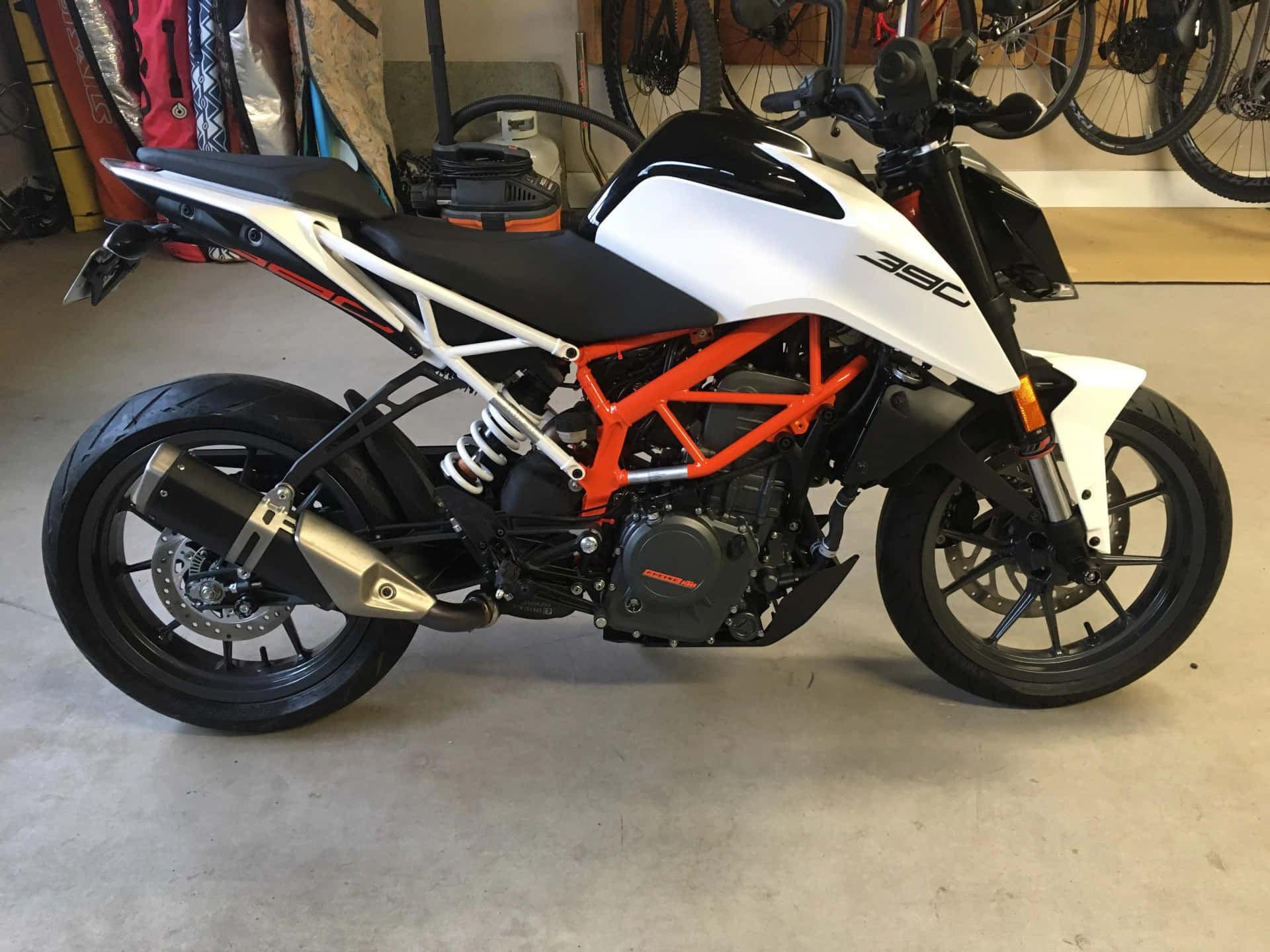  壁紙 — 一輛白色和橙色的KTM 390摩托車停放在室內混凝土地板上，背景掛著自行車。