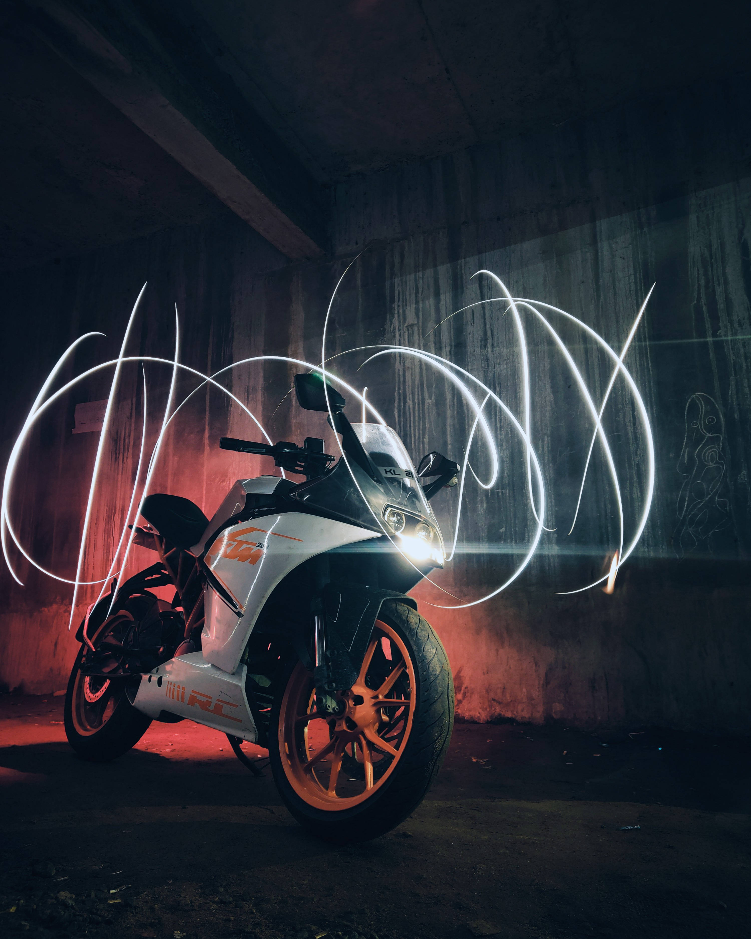 Ktm Rc 390 桌布 壁紙 — 一輛白色和橙色的KTM RC摩托車在粗糙的混凝土隧道中，在戲劇性的紅色燈光和旋轉的白色光軌下閃閃發光。