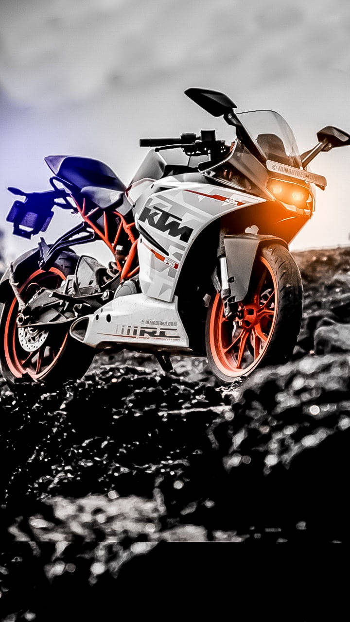 KTM 摩托車 桌布 壁紙 — 一輛白色和橙色的KTM摩托車在陰天之下，亮著車頭燈，矗立在崎嶇的黑色熔岩地上。
