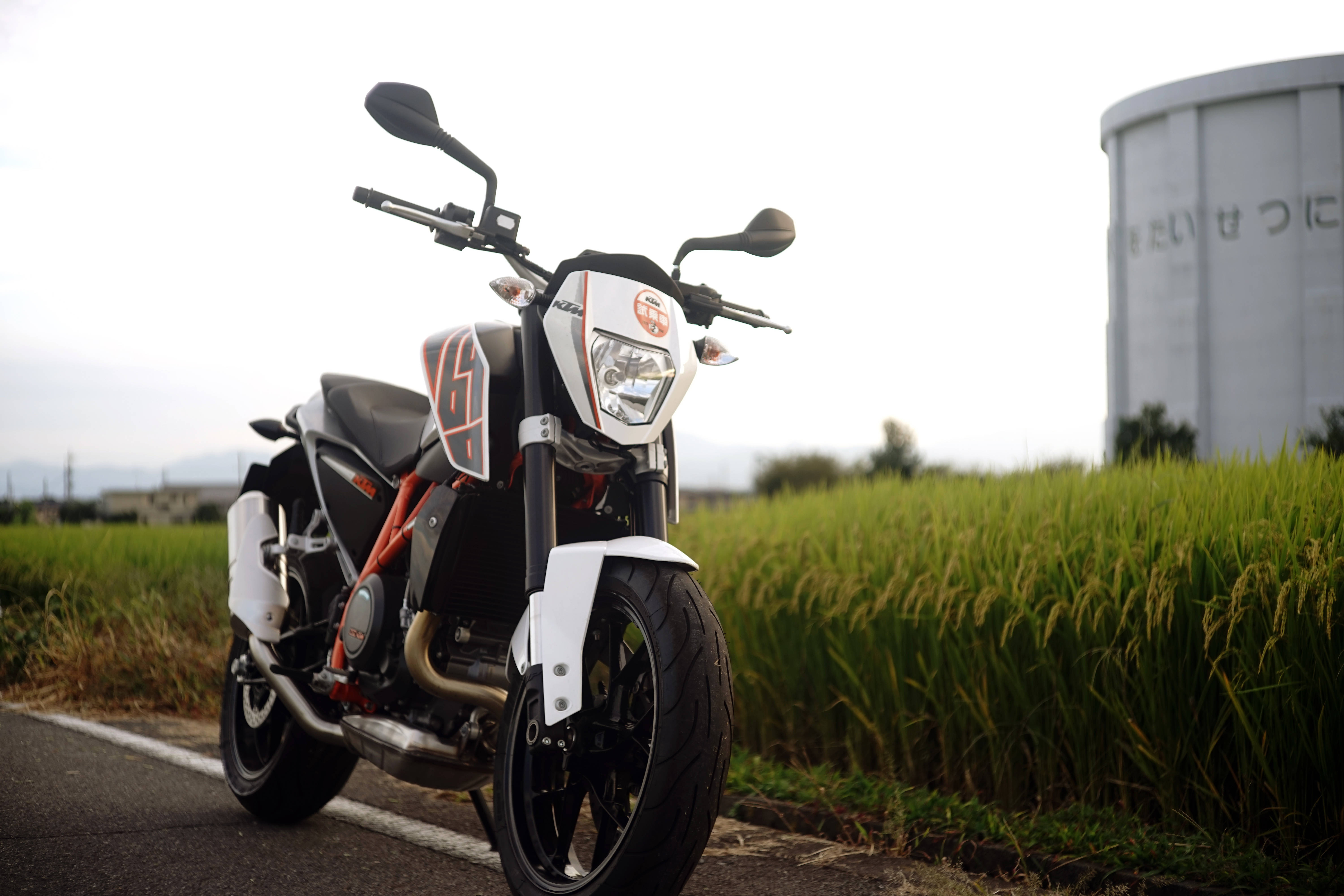 KTM 摩托車 桌布 壁紙 — 一輛白色和橙色的運動機車停泊在鄉村道路上，旁邊是茂密的綠色稻田，天空多雲。