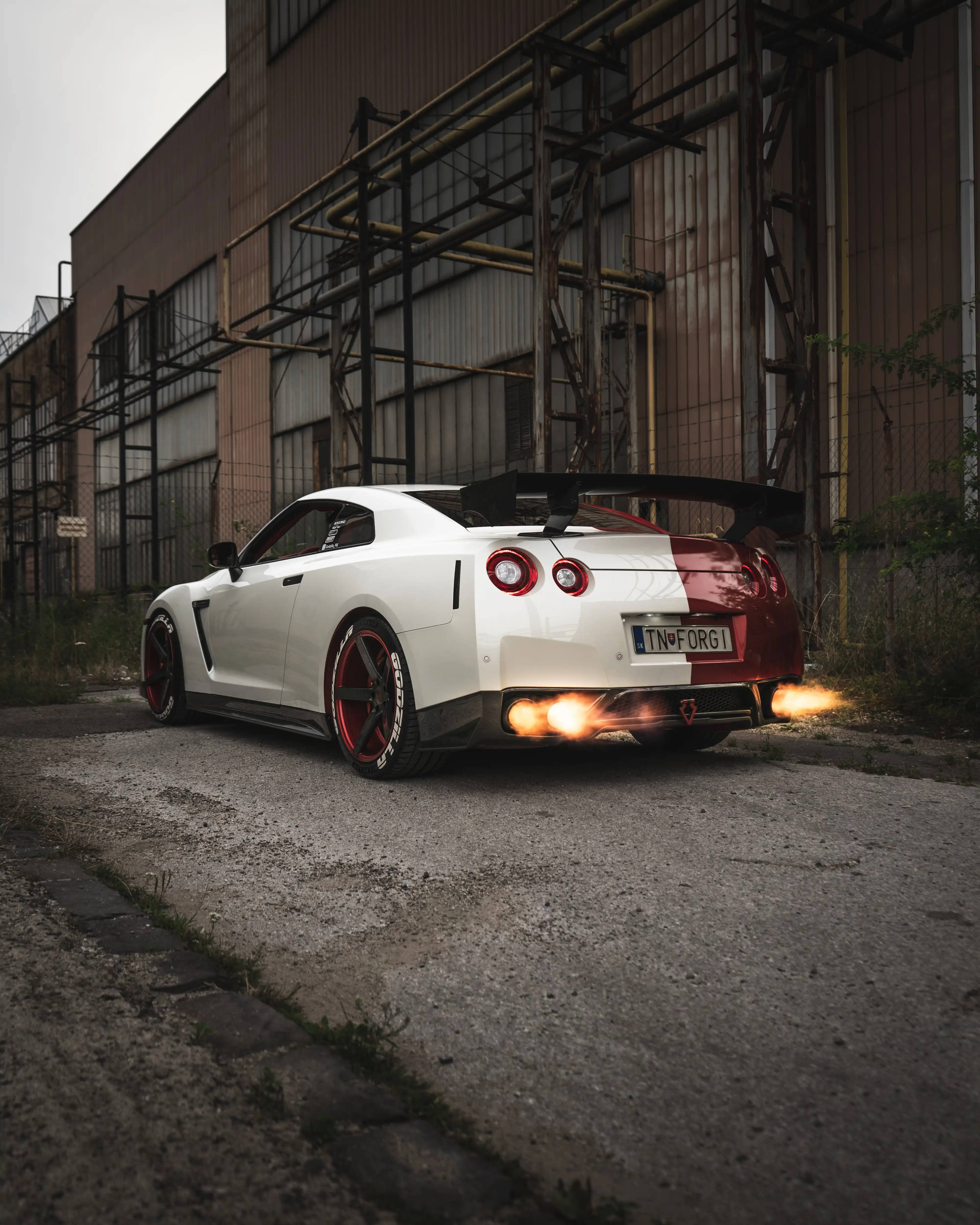 Nissan 桌布 壁紙 — 一輛白色和紅色的日產GT-R，帶有發光的排氣管，停在生鏽的工業建築前。
