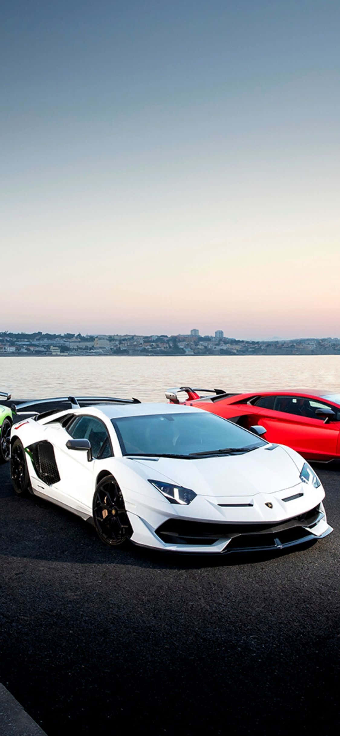 Lamborghini Phone 桌布 壁紙 — 一輛白色和紅色的藍寶堅尼跑車在日落時停泊在海濱，背景是城市天際線。