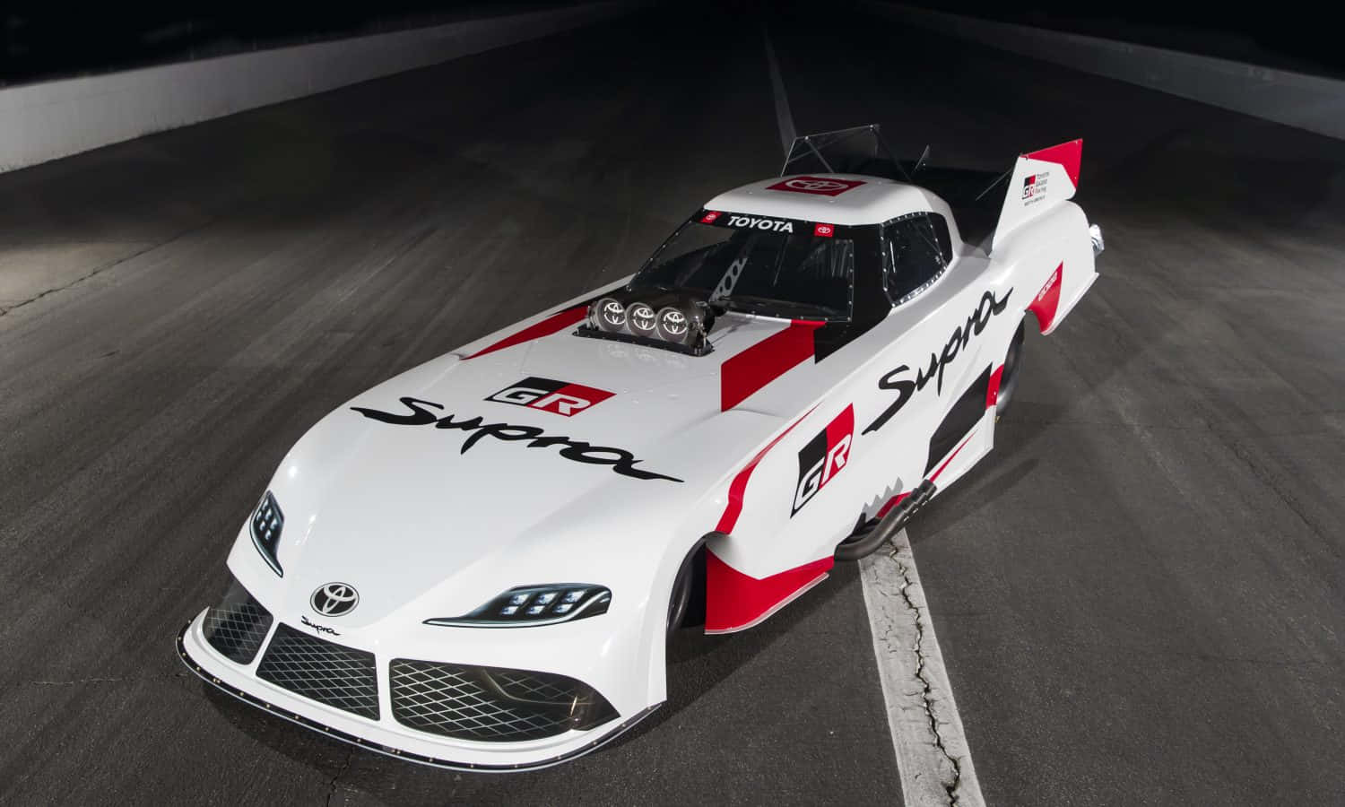 cars 桌布 壁紙 — 一輛白色和紅色的豐田Supra賽車帶有'GR Supra'品牌標誌，在明亮的聚光燈下停泊在黑暗的賽道上。