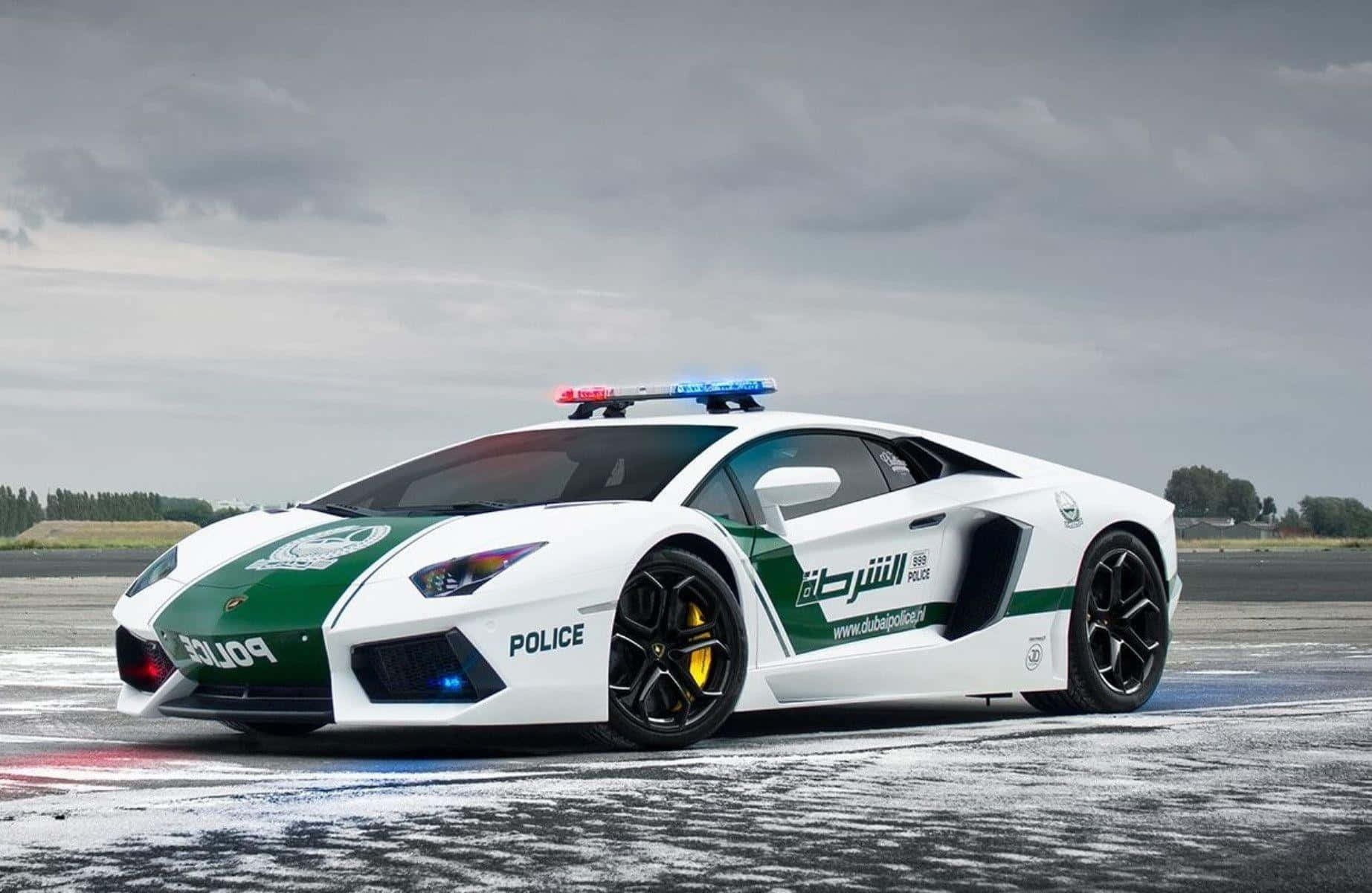Police Car 桌布 壁紙 — 一輛白色和綠色的杜拜警車藍寶堅尼Aventador，在陰天下停在濕瀝青路上，閃著燈。