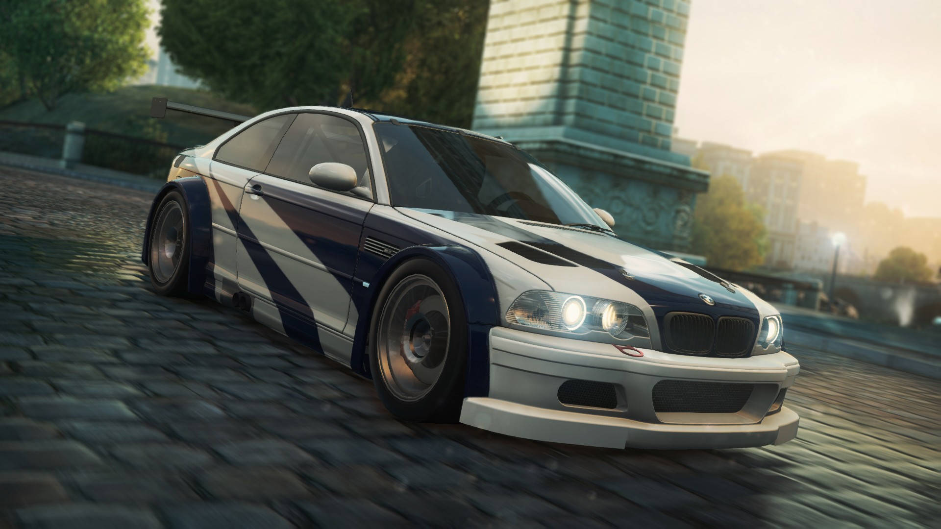 Need For Speed 桌布 壁紙 — 一輛白色和藍色的BMW M3賽車在鵝卵石街道上奔馳，背景有一座石頭紀念碑。