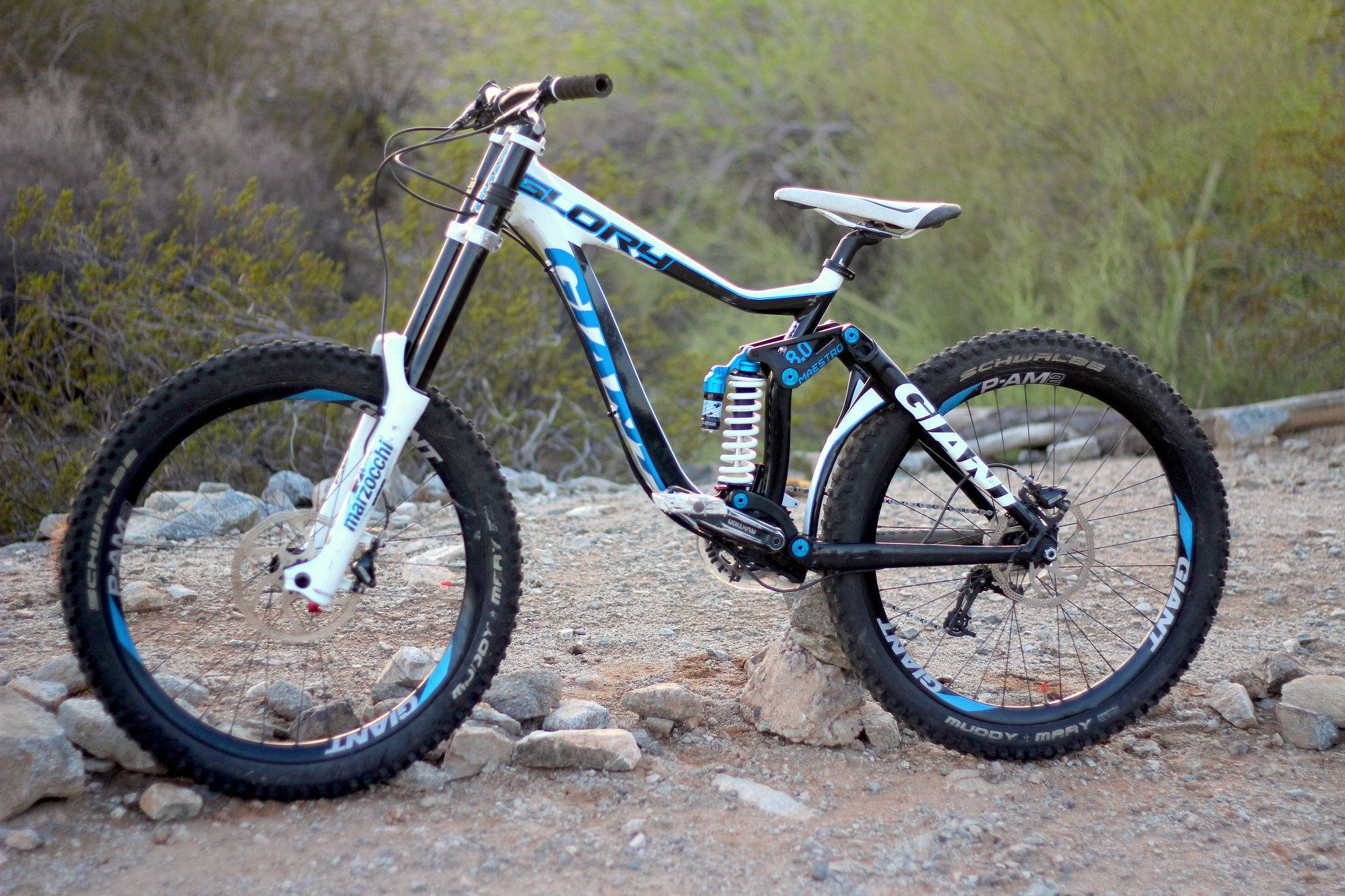 Mountain Bike 桌布 壁紙 — 一輛白色和藍色的全避震登山自行車屹立在岩石地形上，背景有乾燥的灌木叢。