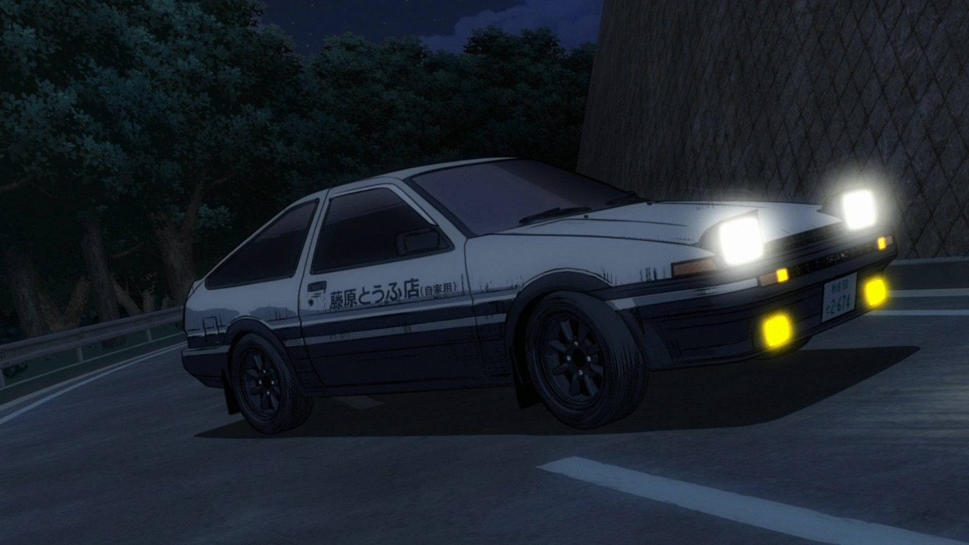 Initial 桌布 壁紙 — 一輛白色和黑色的AE86跑車，車頭燈發光，在夜晚沿著蜿蜒的道路奔馳，道路兩旁有樹木和石牆。