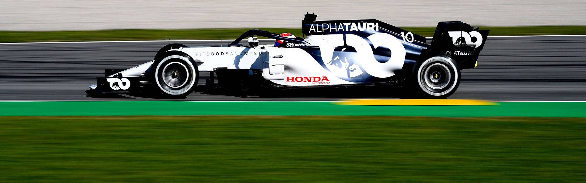 白色 桌布 壁紙 — 一輛白色和黑色的AlphaTauri Honda一級方程式賽車在賽道上疾馳而過，背景是模糊的綠色草地和瀝青路面。