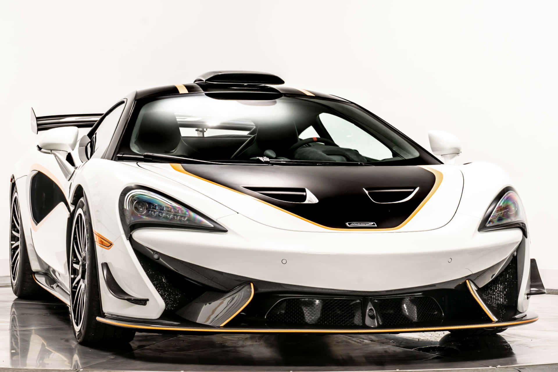 Mclaren 620r 桌布 壁紙 — 一輛白色和黑色的McLaren Senna，帶有金色點綴，在攝影棚燈光下閃耀於反光地板上。
