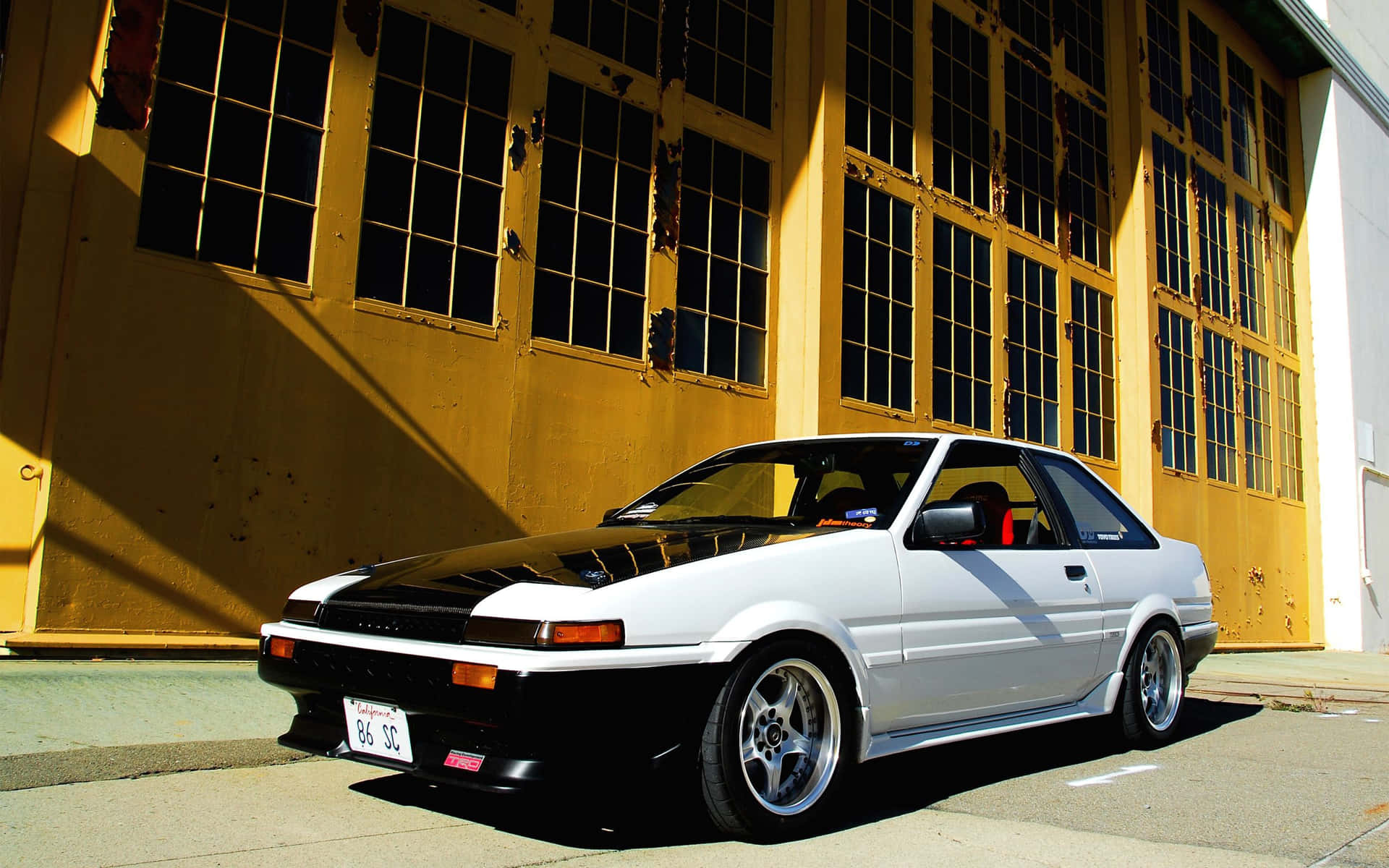 白色和黑色的豐田AE86停放在黃色倉庫前。 — Toyota Supra Mk4 壁紙