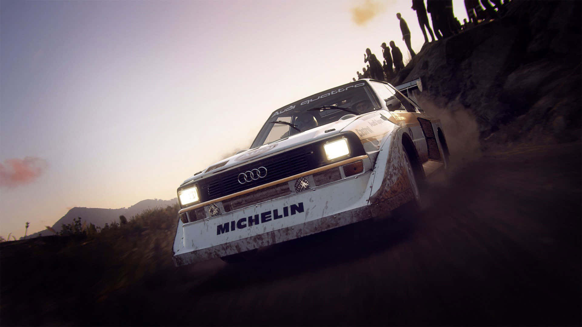 1920x1080 Dirt Rally 桌布 壁紙 — 一輛白色奧迪Quattro拉力賽車在日落時分沿山路疾馳，揚起塵土，觀眾從上方觀看。