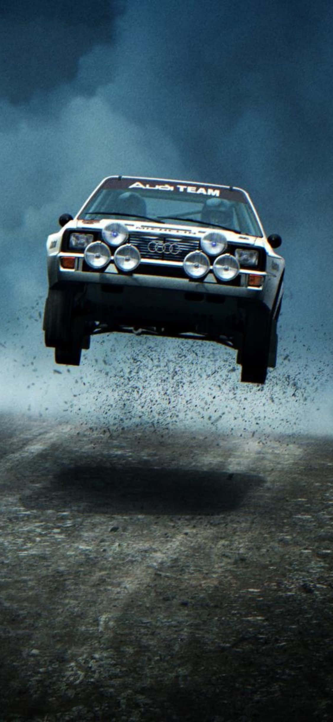iPhone Xs Dirt Rally 桌布 壁紙 — 一輛白色奧迪拉力賽車在暴風雨的天空下，在泥濘賽道上騰空而起，揚起碎石。