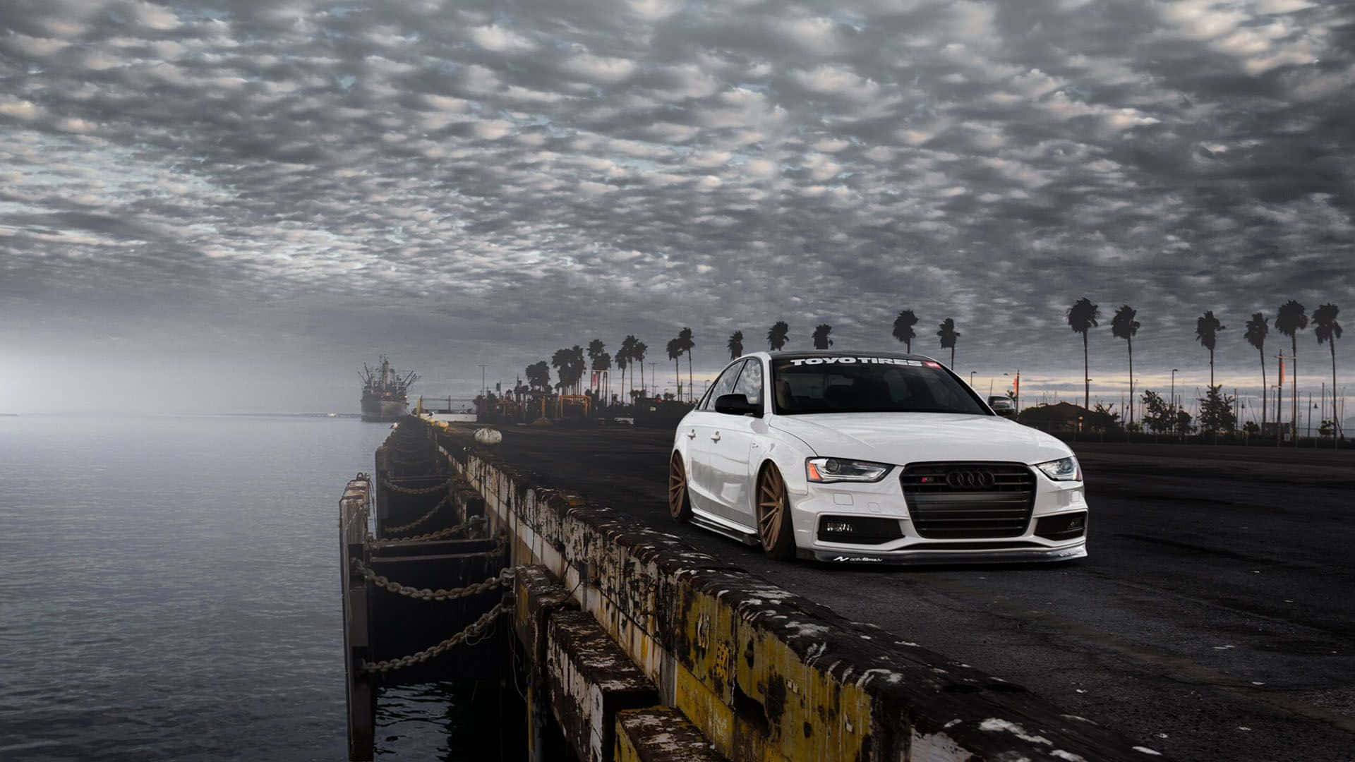Audi S4 桌布 壁紙 — 一輛白色奧迪轎車配金色輪圈，停泊在風化的碼頭上，背景是陰天、棕櫚樹和水面。