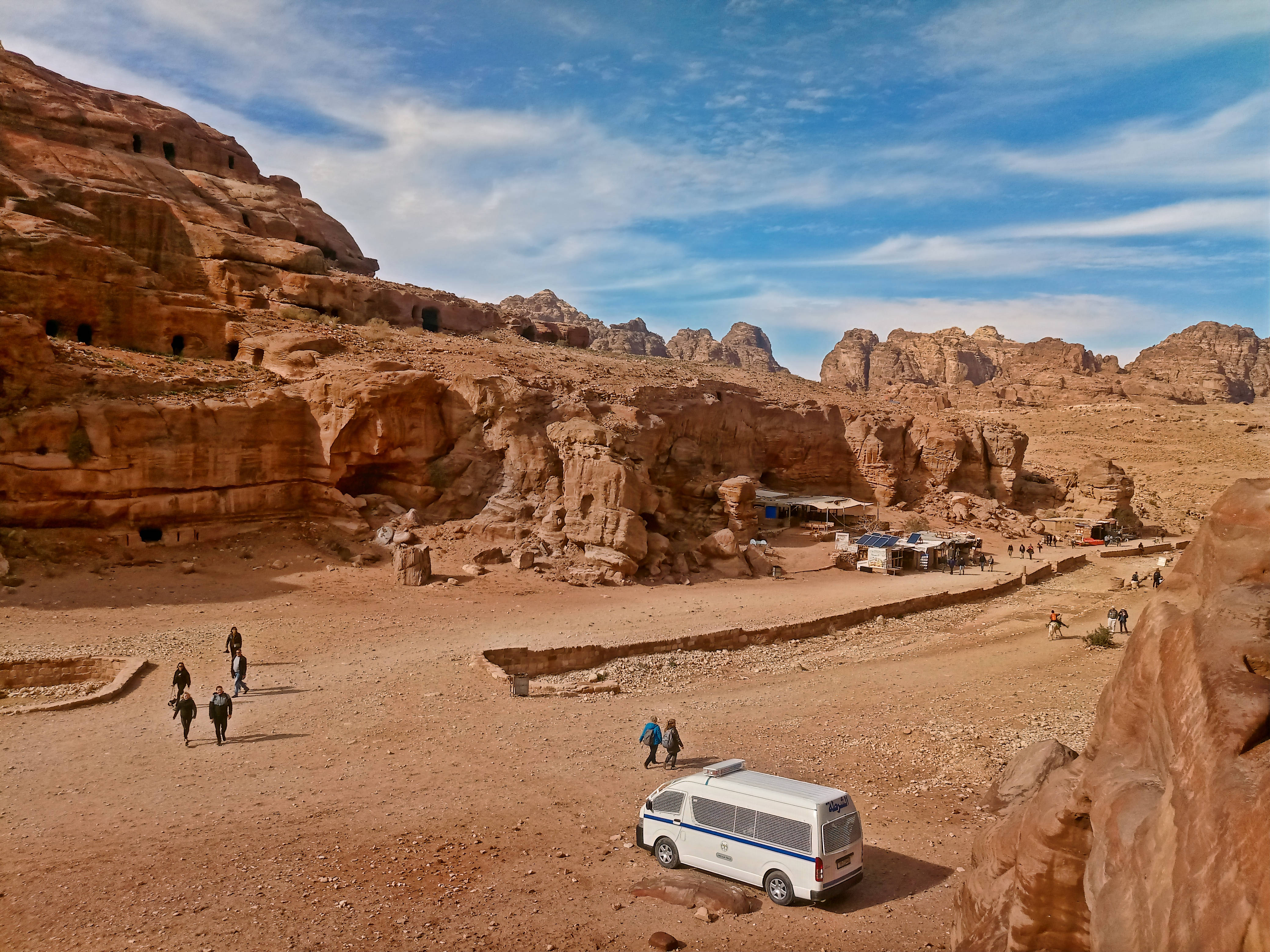 Jordan 桌布 壁紙 — 一輛白色廂型車停放在陽光明媚的沙漠峽谷中，有古老的岩石鑿成的住所和遊客在明亮的藍天下探索崎嶇的紅色砂岩懸崖。