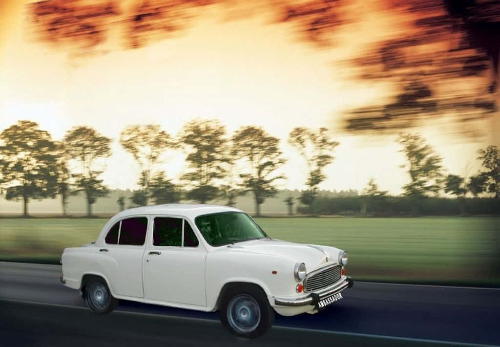 Hindustan Motors 桌布 壁紙 — 一輛白色復古汽車沿著道路疾駛，背景是模糊的樹木和移動中的金色日落天空。