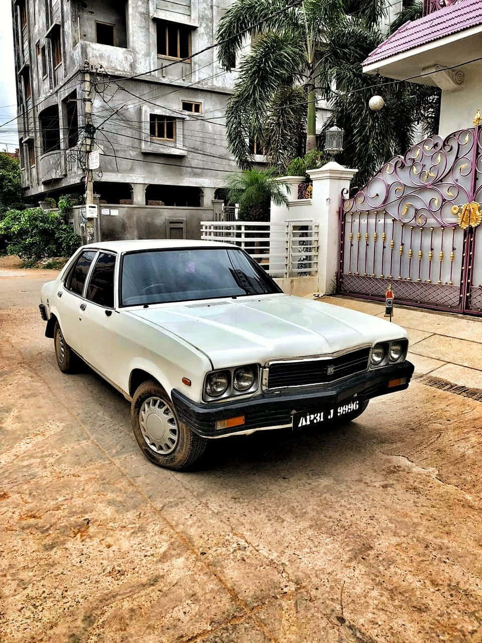 Hindustan Motors 桌布 壁紙 — 一輛白色復古轎車停泊在塵土飛揚的車道上，旁邊是華麗的大門和未完成的混凝土建築。