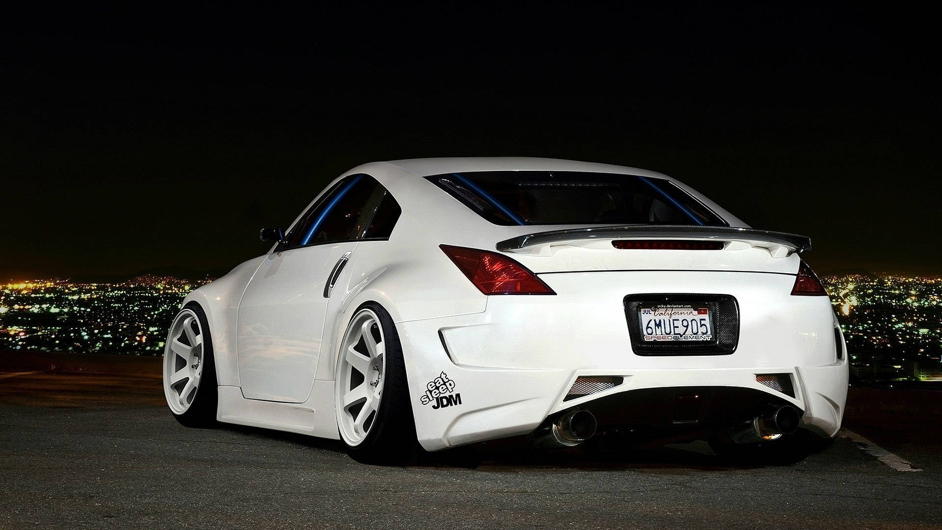 Nissan 350z 桌布 壁紙 — 一輛白色改裝跑車配備定制輪轂和雙排氣管，在夜間停泊，俯瞰著閃爍的城市景觀。