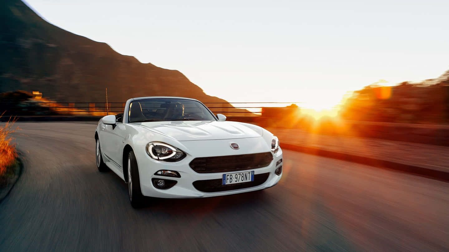 Fiat 124 Spider 桌布 壁紙 — 一輛白色敞篷跑車在日落時分沿著蜿蜒的山路疾馳，身後閃耀著金色的光芒。