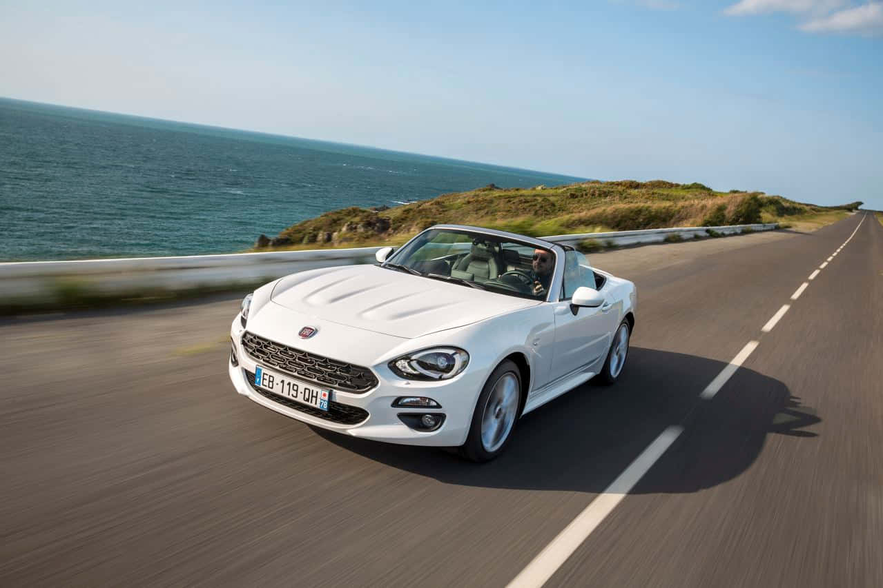 Fiat 124 Spider 桌布 壁紙 — 一輛白色敞篷跑車沿著海岸道路行駛，海洋在晴朗的藍天下延伸至地平線。